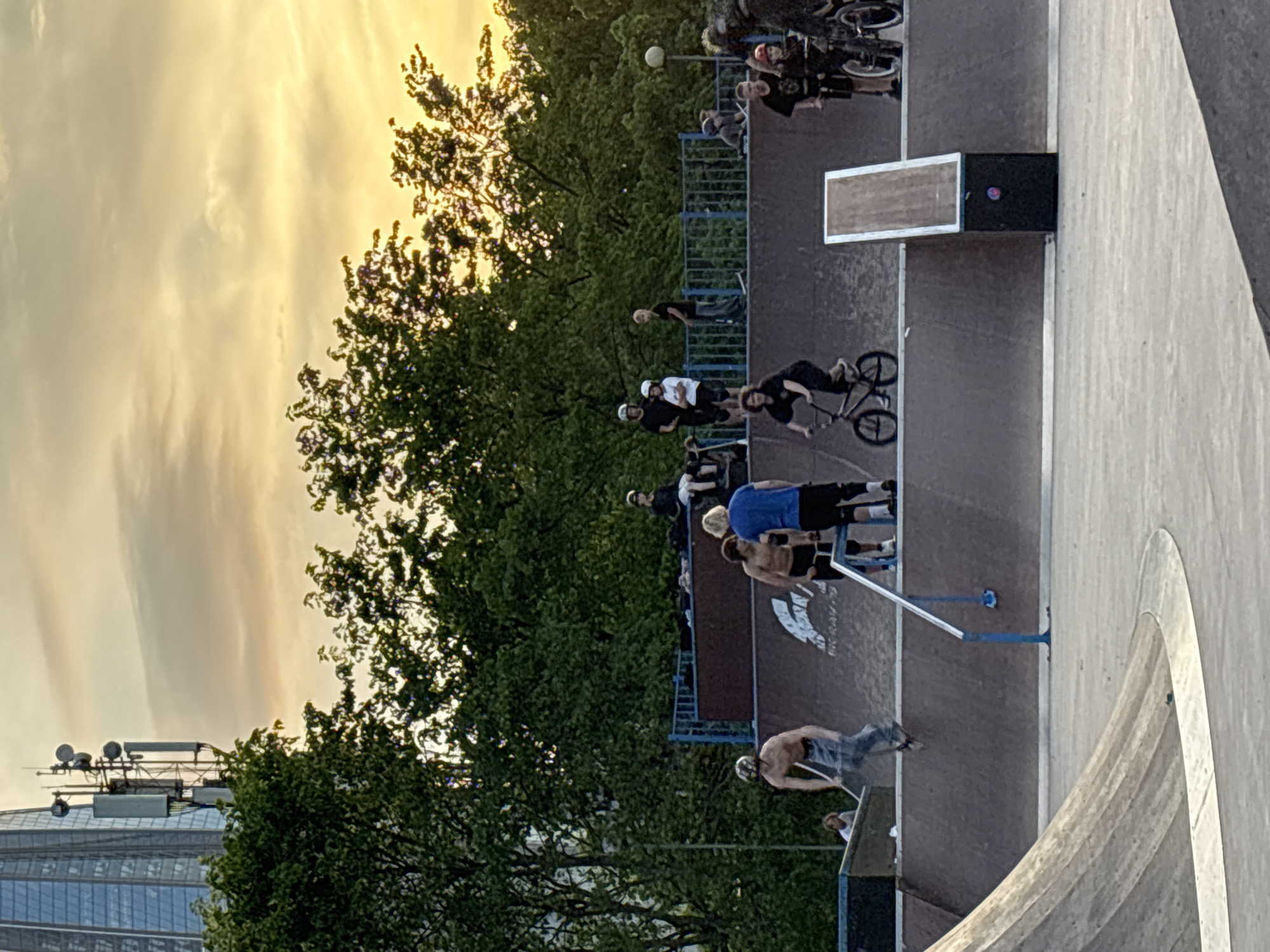 The image depicts an active outdoor skatepark scene during the golden hour, characterized by a warm, yellowish sky with scattered clouds, indicating late afternoon or early evening. A diverse group of people, primarily young adults, are present.
Several individuals are engaged in action sports: multiple people are on BMX bikes, either actively riding or standing with their equipment, and at least one person is seen with a scooter on a blue and white metal grind rail. Others are spectators or resting, standing or sitting around the various ramps and ledges of the park.
The skatepark features a variety of elements, including concrete quarter pipes, a large, flowing concrete bowl in the foreground, and a section with wooden ramps and ledges. A distinct rectangular box/ledge is visible on a darker brown ramp surface. Beneath this, faint white text on the ramp surface appears to read "RK RAILS".
Lush green trees line the perimeter of the skatepark, suggesting a park-like setting. In the distant background on the left, part of a tall, modern building with several communication antennae on its roof can be seen, hinting at an urban environment. The overall mood is casual and social, with individuals enjoying the outdoor activity in clear weather. The location is Saint Petersburg, Russia.