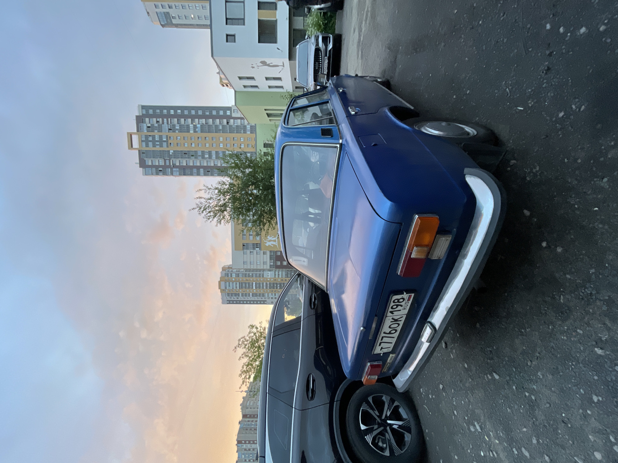 The image depicts a street scene with several cars parked in front of residential buildings. The primary focus is on a blue vintage car, likely from the Soviet era, with a chrome bumper and a visible license plate that reads "T776OK 198 RUS". To the right of the blue car, a newer, dark-colored SUV is parked. In the background, multiple modern apartment buildings of varying heights are visible, suggesting an urban or suburban environment. One building has a white facade with green accents and a mural of a figure. The sky shows a mix of light blue and soft orange hues, indicating that it is either early morning or late evening, possibly during sunset or sunrise. The weather appears to be clear, with a few wispy clouds. The scene is static, with no apparent activity or people. The lighting suggests a time of day when shadows would be elongated.