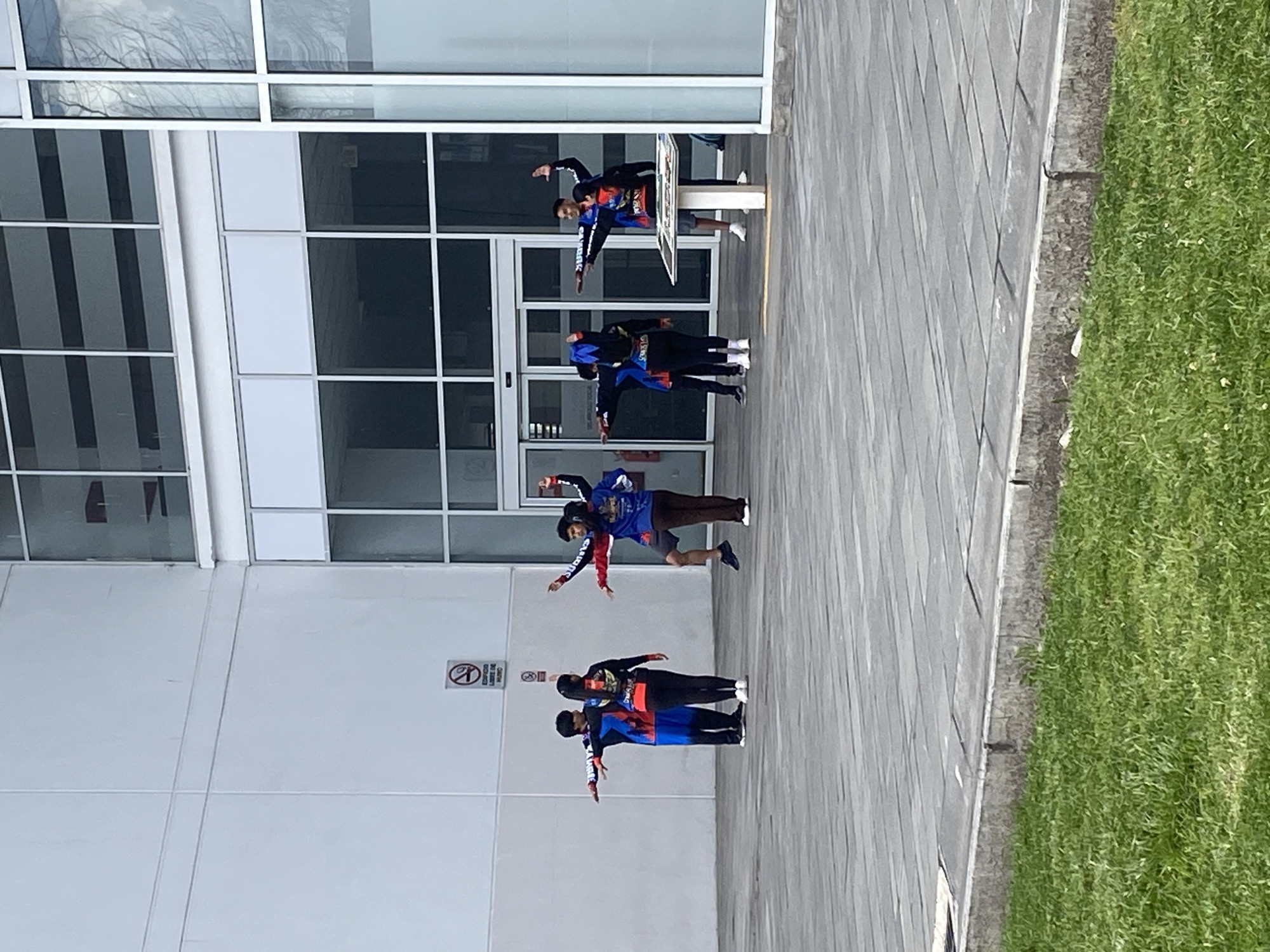 A group of four pairs of people is posed outside a modern building. The building has white walls and large windows. The people wear matching jackets, some in blue, red and black colors, and they stand with their arms extended sideways. A no-smoking sign is visible on the wall next to the pairs of people. The foreground consists of a paved area, with a strip of green grass on the right. The location is likely in Quito, Ecuador.