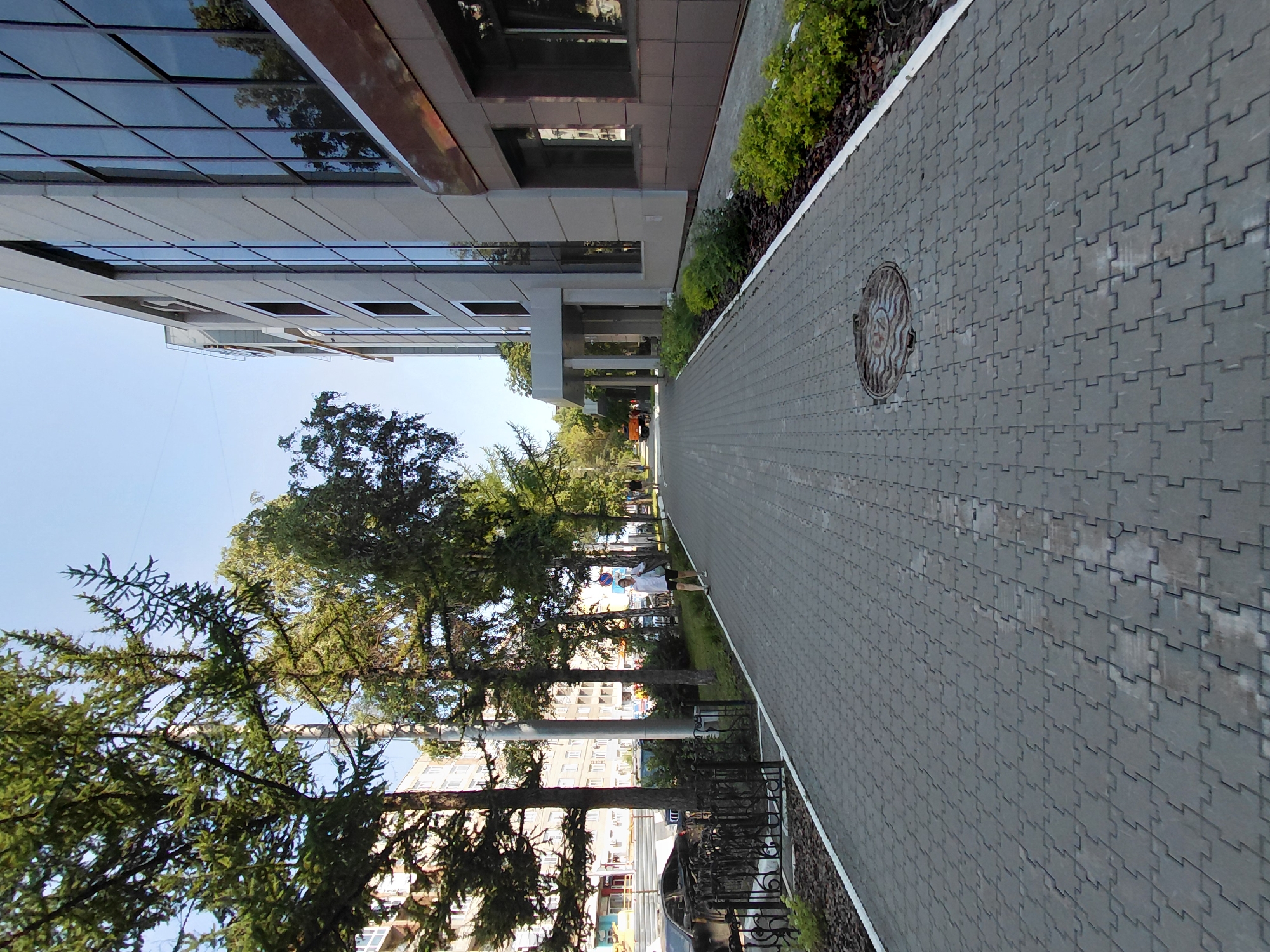 The image shows a paved walkway in Novosibirsk, Russia, in front of a modern building with large glass windows and a light-colored facade. Several trees line the walkway on the left side, casting shadows. A few people are visible walking on the path. In the background, there is a street with some parked cars and what appears to be a building with multiple stories. A man in a white shirt and dark shorts is walking towards the viewer on the right side of the walkway, carrying a bag. A street sweeper or maintenance vehicle is visible in the distance on the street. The sky is clear and blue.