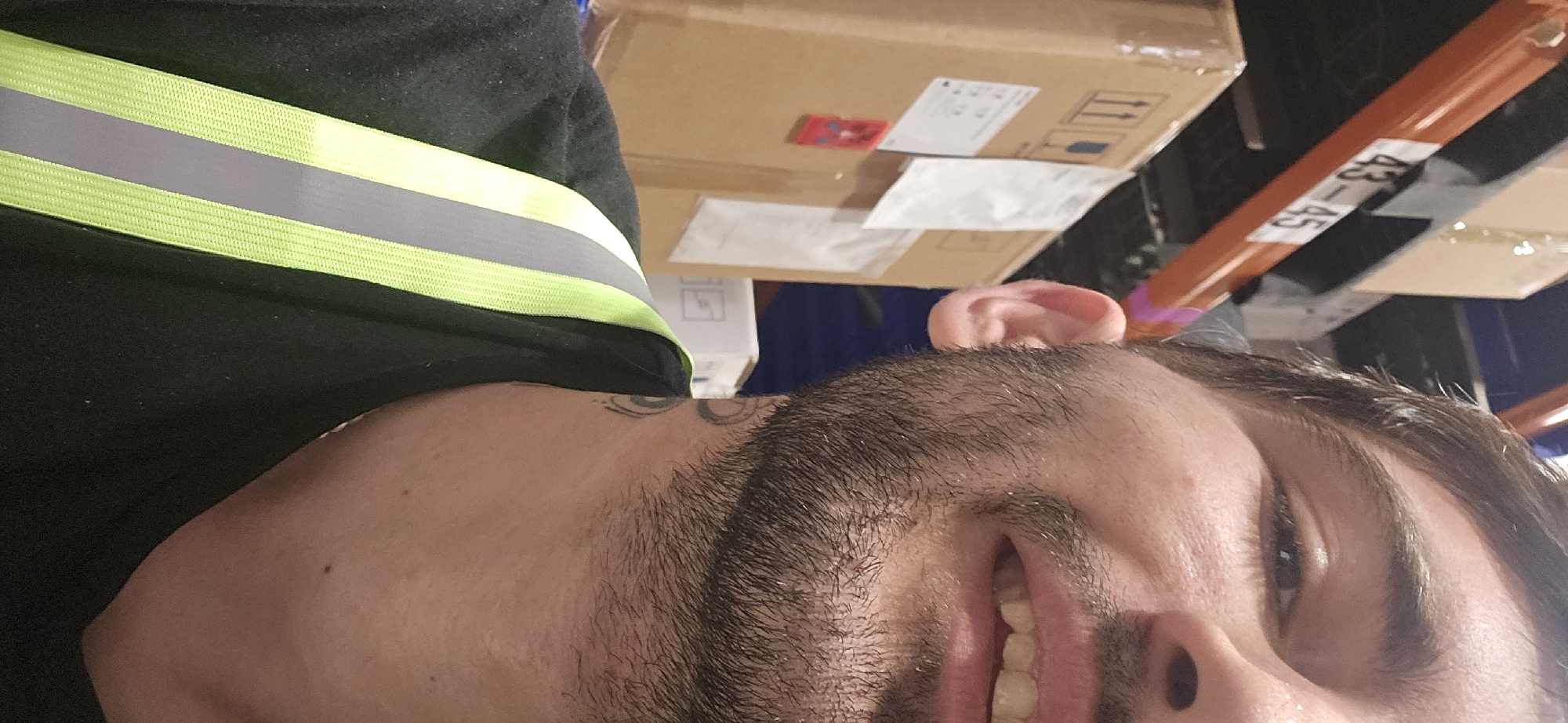 The image shows a man from the chest up wearing a black shirt with a green and gray reflective safety vest. The background includes cardboard boxes, possibly in a warehouse, with an orange rack numbered "43-45". The location is unknown.