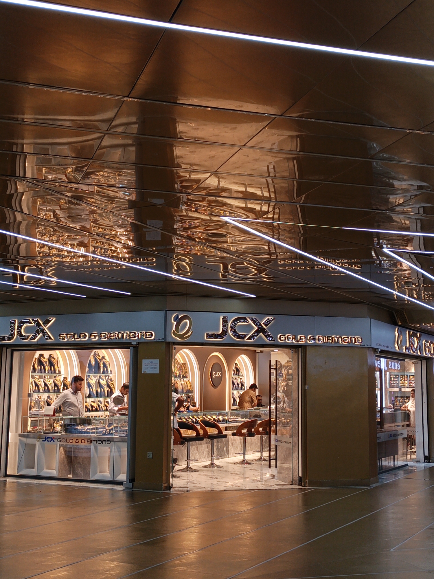 An interior shot of a jewelry store, likely in a mall or covered walkway, features a prominent "JCX GOLD & DIAMOND" sign. The store's facade is a mix of glass display cases filled with jewelry and a golden-colored metallic paneling. Two male employees are visible behind the counter in the left section of the store, one in a light-colored collared shirt and the other in a beige shirt. They appear to be attending to customers or arranging merchandise. In the right section of the store, a female employee in a white top is seen. The ceiling of the area is made of reflective, gold-toned panels with linear lighting fixtures. The floor is tiled and reflects the overhead lighting and store displays. The overall scene conveys a luxurious and well-lit retail environment.