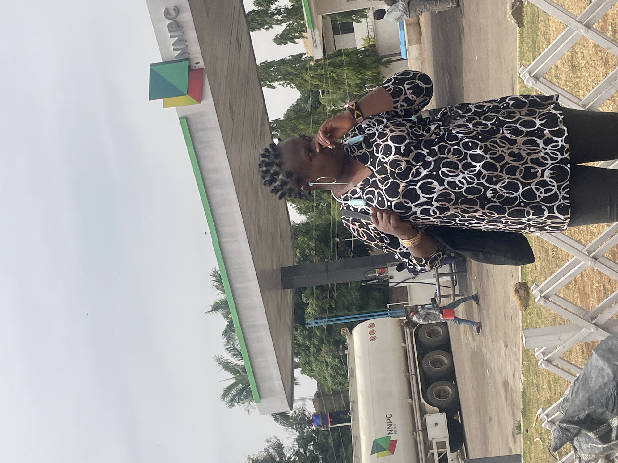 The image shows a woman standing outside an NNPC (Nigerian National Petroleum Corporation) petrol station. She is wearing a black and white patterned blouse and black trousers. She has her hand near her face. A fuel tanker truck with the NNPC logo is parked nearby. In the background, there is a building and trees. The image was taken outside, likely during daylight hours. The exact location in Jos, Nigeria, cannot be determined from the image.