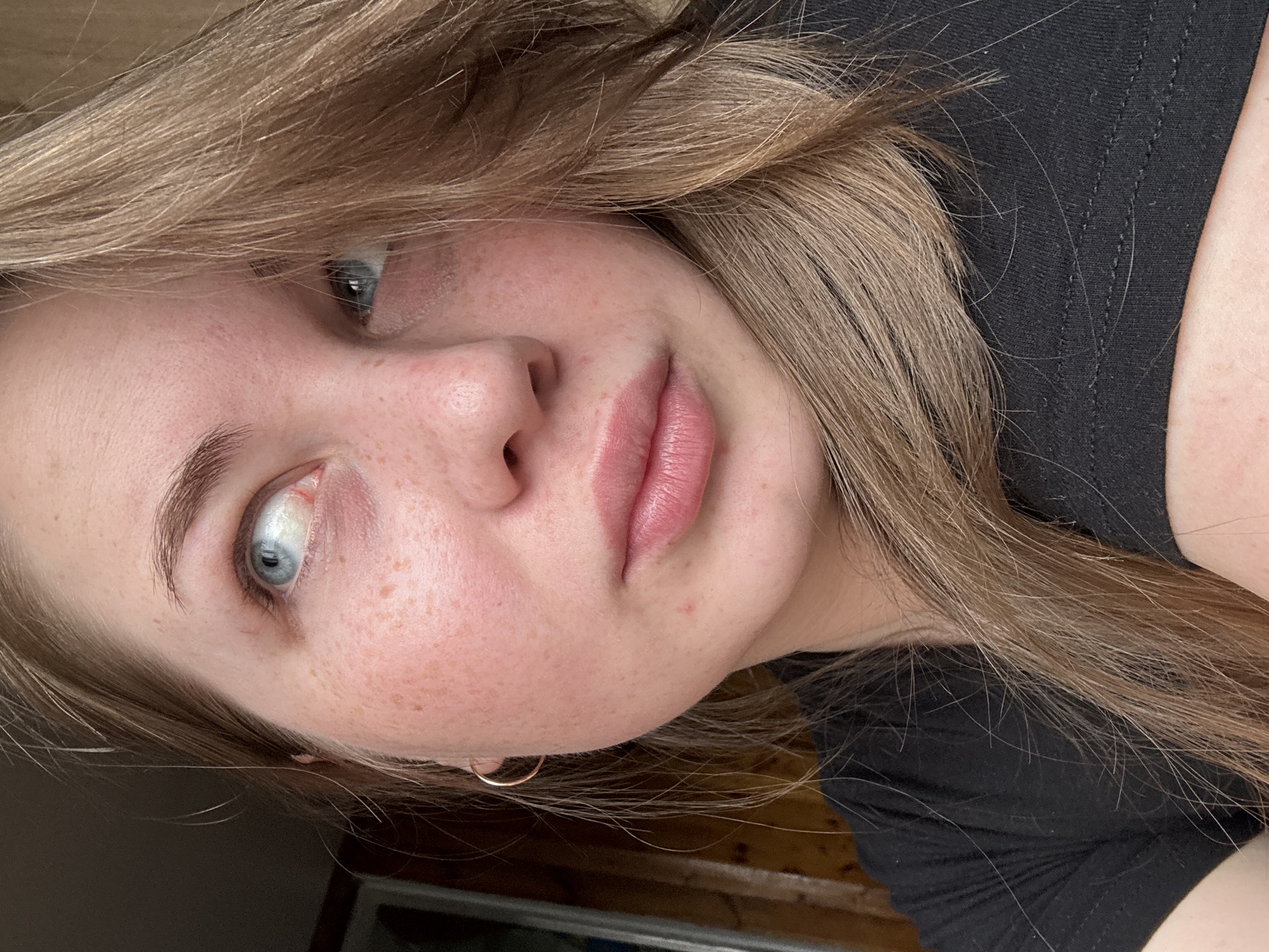 A close-up, rotated selfie of a young, fair-skinned female with light brown, layered hair. Her hair is slightly disheveled and falls across her forehead and cheek. She has light blue eyes and noticeable freckles on her face. Her lips are pink and full. She is wearing a black t-shirt and a small, gold hoop earring in her left ear. The background appears to be a wooden surface, possibly a table or door. There is no information to confirm the location is Уральское, Russia.