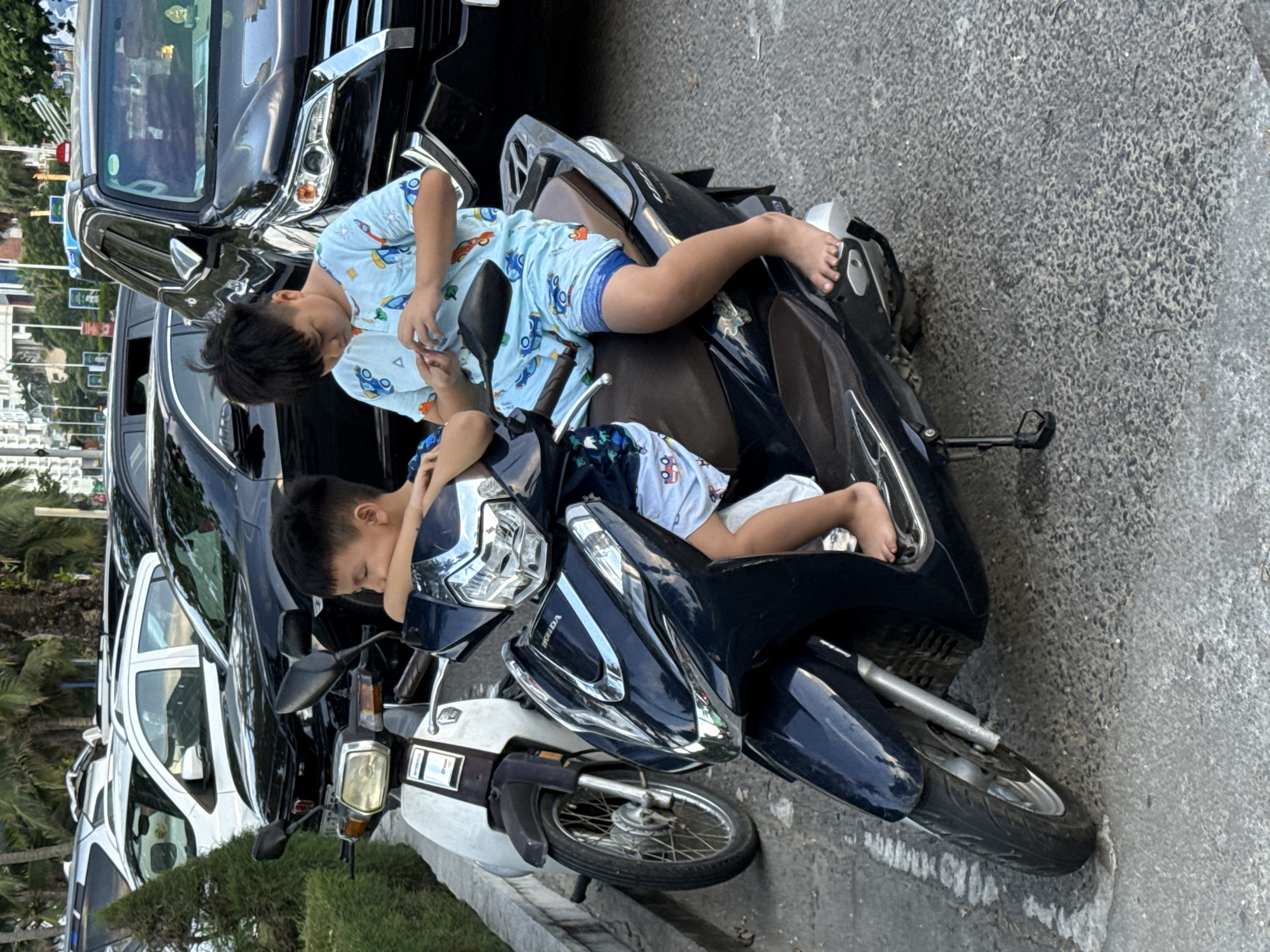 Two young boys are resting on a dark blue and white scooter parked on a paved street in Nha Trang Ward, Vietnam. The boy on top is wearing a light blue shirt with a cartoon pattern of cars and trucks, and patterned blue shorts. He is leaning back against the seat, his head resting on his arm. The boy underneath him is wearing a similar patterned shirt and shorts, and is lying down with his head on the seat. His legs are outstretched. Both boys appear to be relaxed.
Behind the scooter, several dark-colored cars are parked. One car, a black SUV, is particularly prominent. In the background, there are palm trees and some buildings, suggesting a tropical or coastal environment. The ground is paved with asphalt and has some white markings. The lighting suggests it is daytime, likely outdoors. There is no visible text in the image.