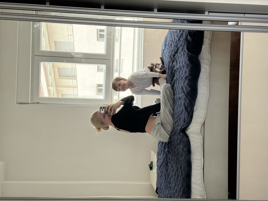 The image displays a mirror selfie reflecting an indoor residential scene in Budva, Montenegro. An adult female and a young child are depicted on a bed. The adult female, appearing to be in her 20s or 30s with blonde hair in a bun, is seated on the bed, facing away from the mirror with her head turned back. She is wearing a black long-sleeved crop top and light grey sweatpants, holding a smartphone to capture the reflection, and is smiling. A young male child, approximately 3-6 years old with light hair, is kneeling on the bed behind and to the right of the female, facing the mirror and smiling broadly. He is dressed in a white long-sleeved shirt and brown patterned pants; a small brown stuffed animal is visible near his right foot. The bed features a white mattress and a dark blue, textured blanket. To the left of the bed, a white nightstand holds a small green item. The room has plain white walls. A large window with white frames behind the bed offers an external view of light-colored residential buildings featuring multiple windows with roller shutters and balconies, consistent with urban architecture in Budva. The reflection is captured in a full-length mirror, likely part of a wardrobe, evidenced by a silver vertical frame on the right.