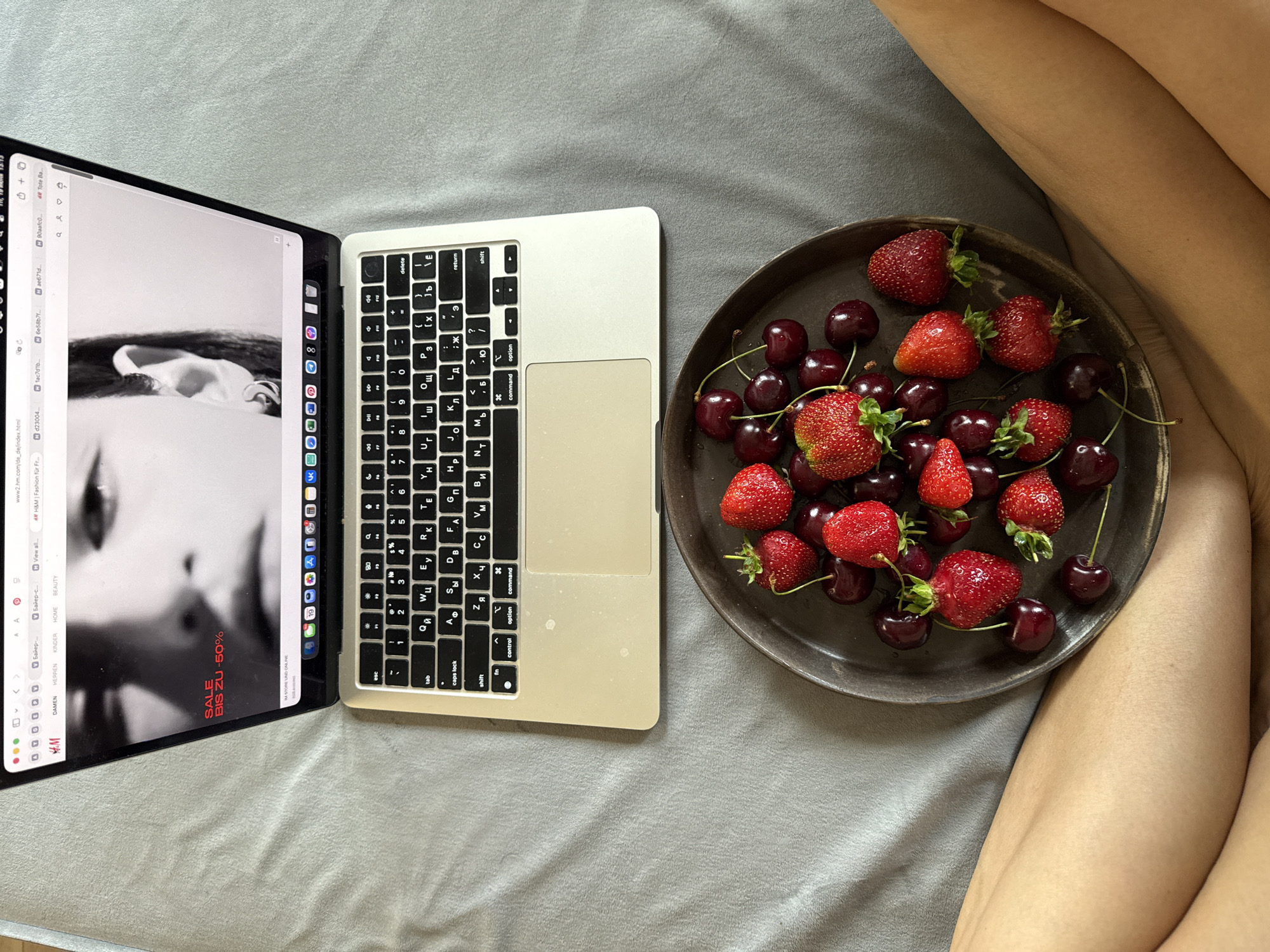 A close-up overhead shot shows a laptop open to a website displaying a fashion advertisement with a black and white image of a person's face. The laptop keyboard is visible, featuring both Latin and Cyrillic characters. To the right of the laptop, a dark, round plate is filled with fresh strawberries and cherries. The scene is set on a grey, textured surface, likely a bed or sofa, with a person's legs visible on the right side of the frame, their skin toned and smooth. The lighting suggests a daytime setting, possibly indoors, with soft shadows indicating natural light. The overall mood is relaxed and casual, hinting at a moment of leisure or work. Visible text on the laptop screen includes "www2.hm.com/de_de/index.html," "H&M | Fashion für Fr...," and "SALE BIS ZU 50%," indicating the website is for H&M and a sale is in progress. The keyboard's Cyrillic characters and the presence of legs suggest this image may be from Chaikovsky, Russia.