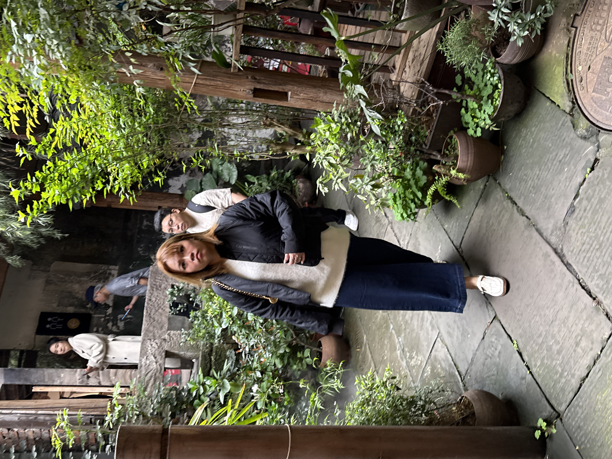 The scene depicts a pedestrian pathway within a densely vegetated, multi-level urban environment identified as Yuzhong District, Chongqing, China, confirmed by a visible manhole cover inscribed "CHONGQING MOUNTAIN CITY."
Four individuals are present. In the immediate foreground, a female with light brown hair, wearing a black bomber jacket over a white knitted top, dark blue wide-leg jeans, and white flat shoes, walks directly towards the viewer with a neutral expression. A thin gold chain, possibly from a shoulder bag, is visible over her right shoulder. Behind her, slightly to the left, a male wearing glasses, a light-colored short-sleeved shirt, and a dark backpack follows, looking forward. On a raised stone platform to the upper left, a female in a light-colored trench coat stands holding a mobile phone in both hands, looking left. Behind her, a male in a grey t-shirt and a dark baseball cap worn backward is visible from the waist up, looking down at a mobile phone he holds in his right hand.
The pathway consists of grey, irregularly shaped flagstones that appear damp or worn. Both sides of the path are densely lined with various green plants and shrubs, many housed in terracotta and darker ceramic pots. Taller leafy plants and small trees create a lush, semi-enclosed atmosphere. Exposed wooden pillars or beams are visible, notably a large one on the right side of the path. Rustic stone or concrete structures form the raised platform and background walls. A black sign with white male and female stick figures, indicative of a restroom, is mounted on the wall of the raised platform.