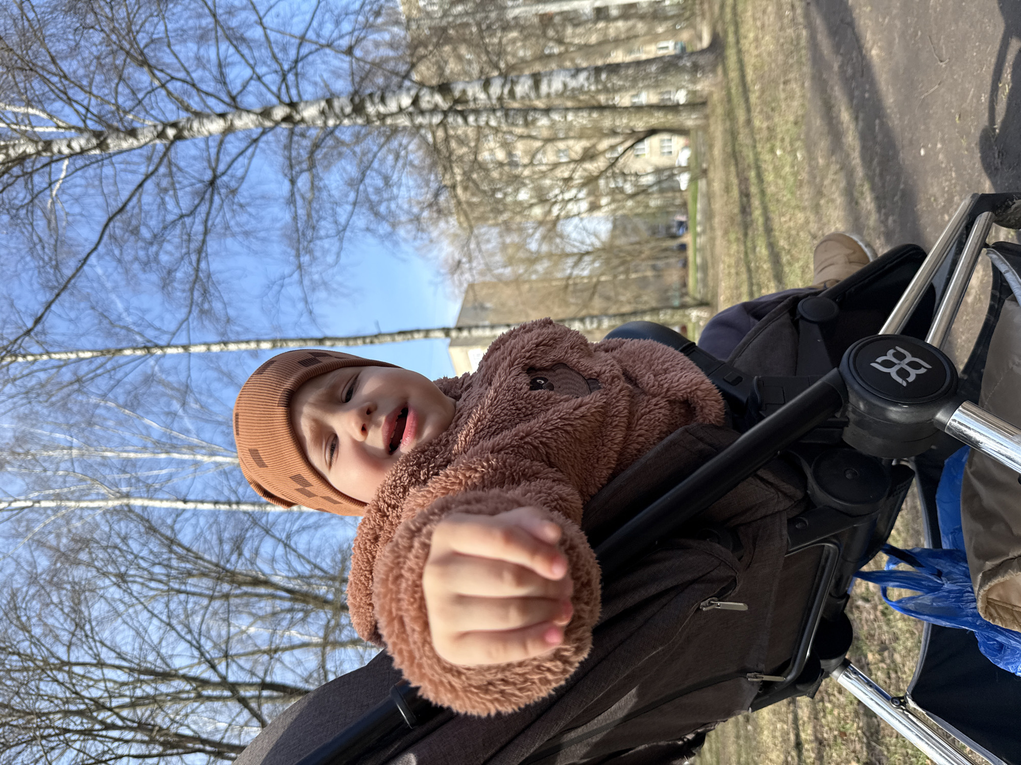 A young child, possibly a toddler, is seated in a stroller outdoors. The child is wearing a brown beanie hat and a brown, fuzzy jacket. The child's left hand is prominently displayed, clenched in a fist. The stroller has a dark-colored fabric seat and frame, with some metallic components visible. In the background, there are leafless trees, a blue sky, and a building in the distance. The environment suggests a park or a residential area on a sunny day.