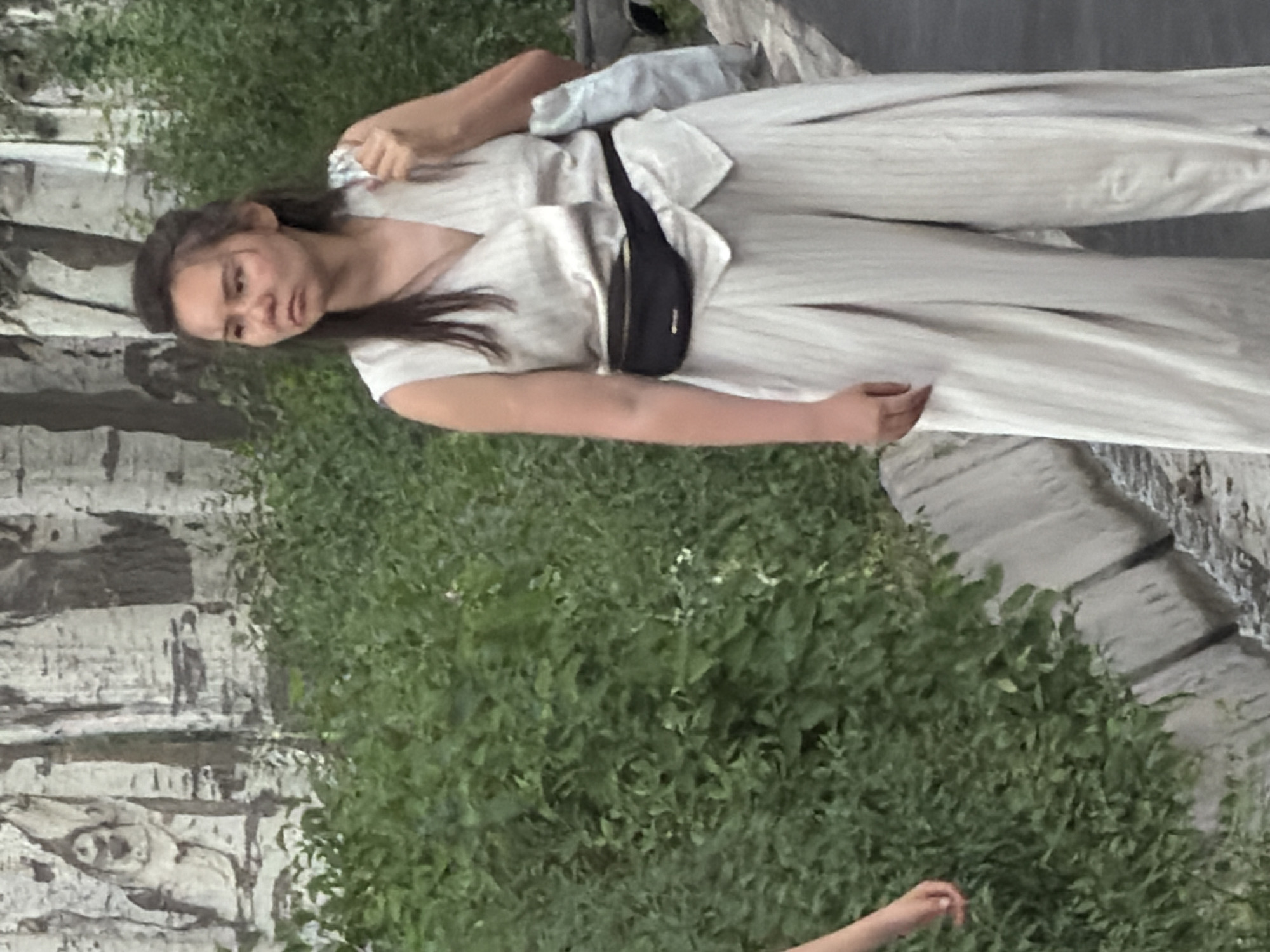 In the city of Unknown, Unknown, a young Asian woman stands outdoors, facing right. She is wearing a sleeveless white vest with a V-neck, a black fanny pack around her waist, and wide-legged white striped pants. Her hair is dark and pulled back in a messy bun. A white bag is slung over her right shoulder. The background consists of green foliage and a light-colored rocky wall. A concrete ditch runs along the bottom of the image.