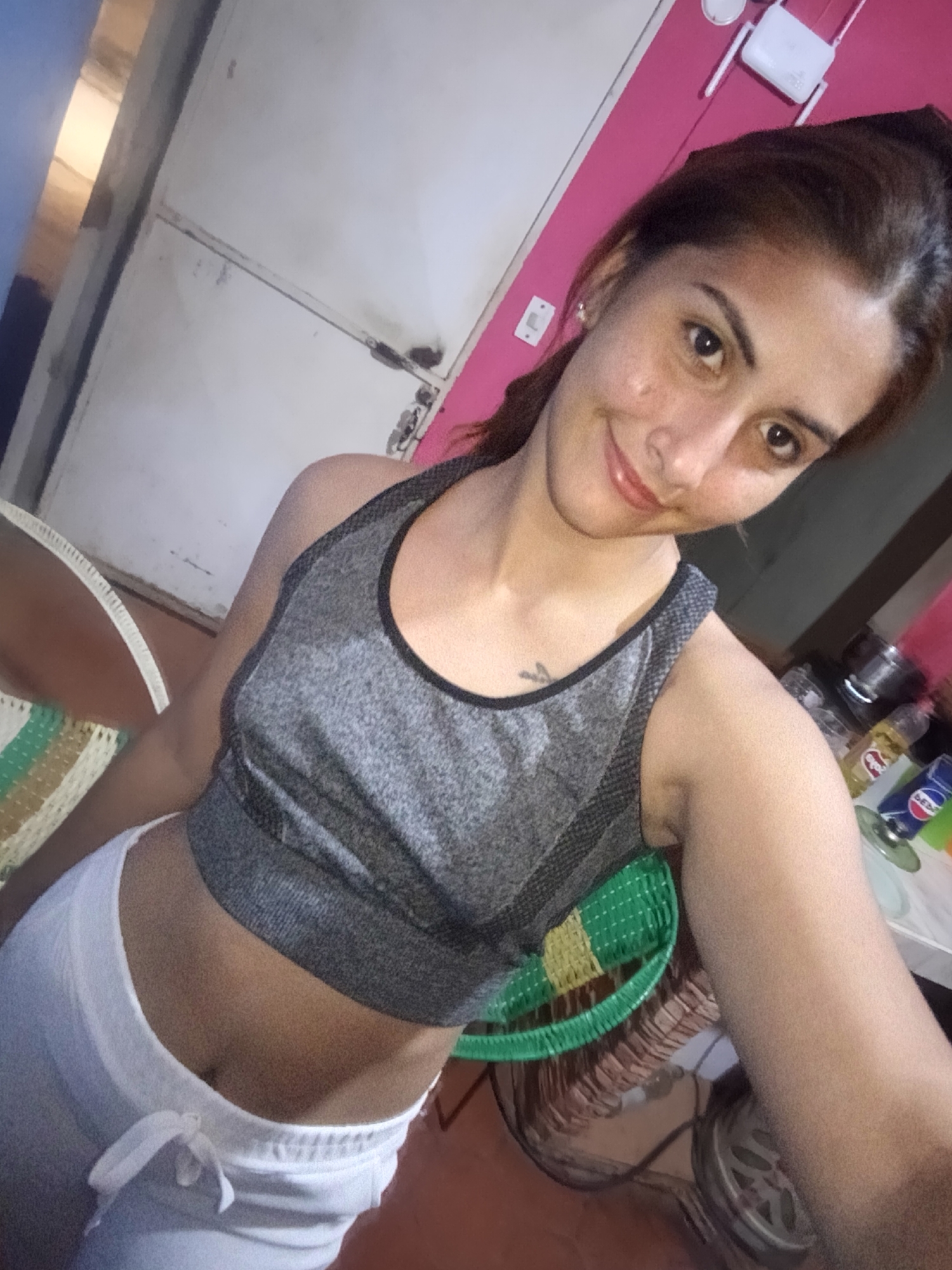 This media file features an indoor selfie of a young woman, likely in her 20s or early 30s. She is positioned slightly to the left, looking directly at the camera with a gentle smile. Her brown hair is pulled back, and she has visible freckles or minor blemishes on her face. She wears a grey athletic crop top or sports bra with black trim, revealing her midriff, and white drawstring sweatpants or shorts. A small, black tattoo is visible on her left upper chest, just above the top.
The setting appears to be a casual home environment, possibly in Juan Griego, Venezuela, as indicated by the context. The background is divided into several sections. To the woman's upper left, a white door or cabinet is visible, showing some wear and possibly dirt. A sliver of a blue wall or surface is on the far left. Behind and to the right of her head, a vibrant pink wall dominates, featuring a white Wi-Fi router with antennas mounted on it, and a white electrical outlet or light switch.
To her lower right, a cluttered surface, resembling a kitchen counter or table, holds various items. These include metal pots or containers, a yellow bottle, and a blue bottle with the partial text "DEXI" visible. Part of a green and yellow woven chair or basket is also seen. On her lower left, another woven chair, in white and green, contributes to the informal atmosphere. The lighting suggests an indoor setting, likely with artificial illumination, indicating it could be evening or night. The woman's expression conveys a sense of comfort and ease.