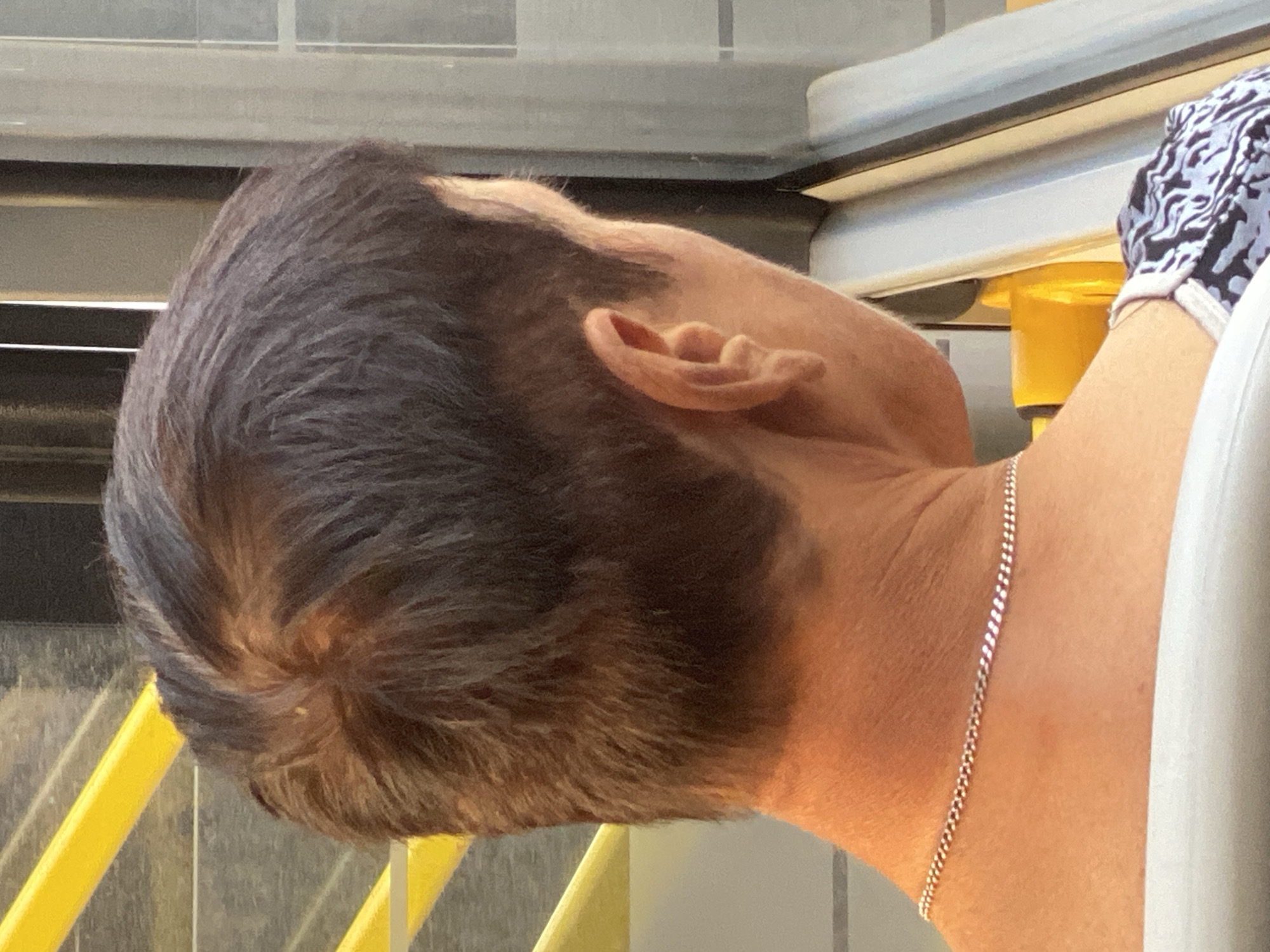 The image is a close-up, rear-view shot of a person's head and neck. The person has short, dark hair with some thinning on top and a shaved or faded style on the sides. A silver chain necklace is visible around their neck. They appear to be sitting on public transport, indicated by the yellow handrail and gray seat in the background, suggesting a location within the city of Novokuznetsk, Russia. The lighting is natural, suggesting daytime.