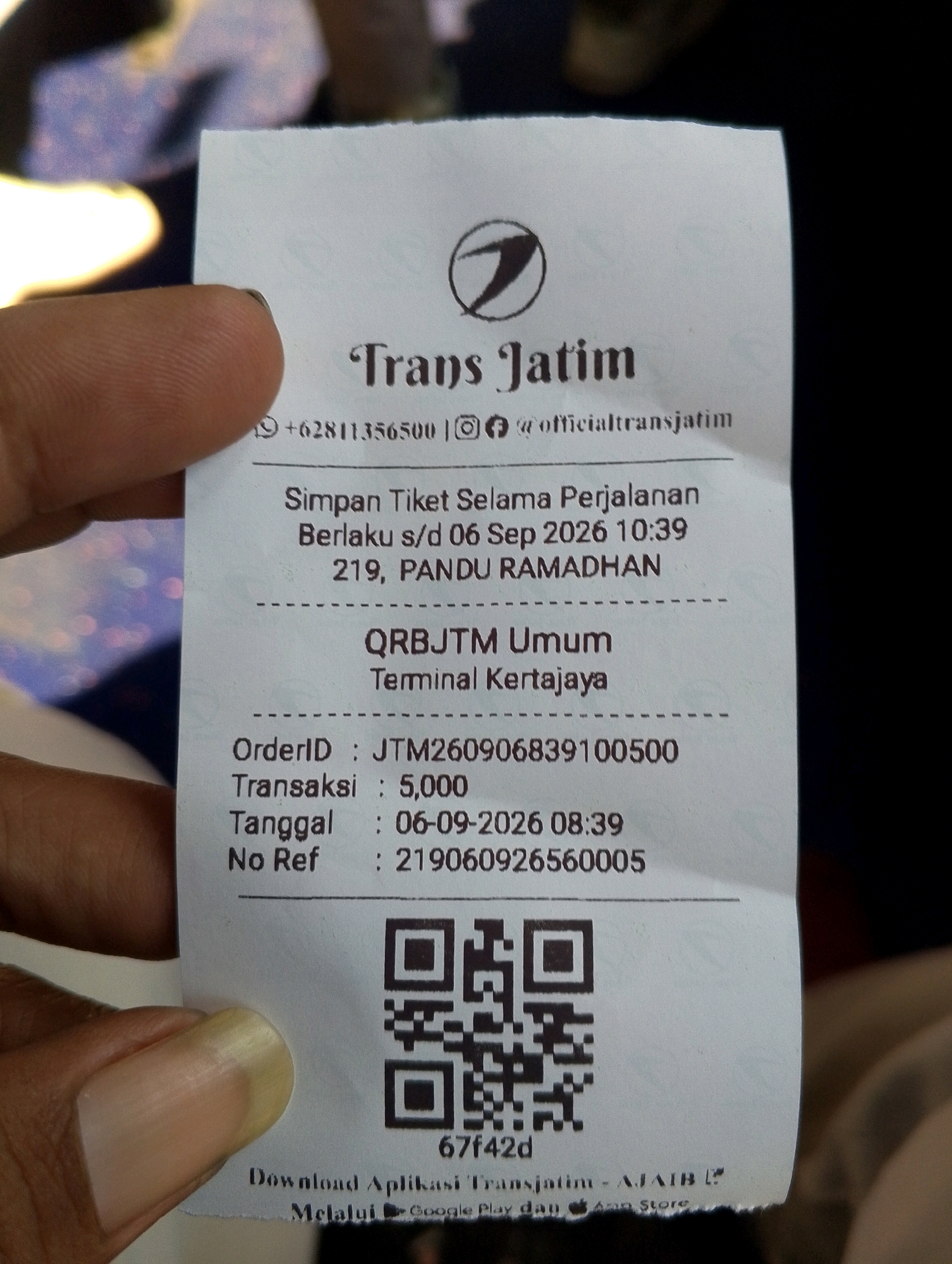 The image displays a close-up view of a Trans Jatim ticket held by a person's hand. The ticket is primarily white with dark text and graphics. At the top, a logo with the text "Trans Jatim" is visible, followed by contact information including a phone number and social media handles. Below this, details about saving the ticket for travel are presented, along with a validity date of "06 Sep 2026 10:39" and the location "219, PANDU RAMADHAN". Further down, information about the ticket is listed, including "QRBJTMI Umum", "Terminal Kertajaya", "OrderID : JTM260906839100500", "Transaksi : 5,000", "Tanggal : 06-09-2026 08:39", and "No Ref : 219060926560005". A QR code is prominently displayed below these details, with the code "67f42d" directly beneath it. The bottom of the ticket shows instructions for downloading the Trans Jatim application via Google Play and the App Store. The background is out of focus, showing a blurred pattern of colors, likely a garment or seating. The overall context suggests a purchased ticket for transportation in or around Gunung Gedangan, Indonesia.