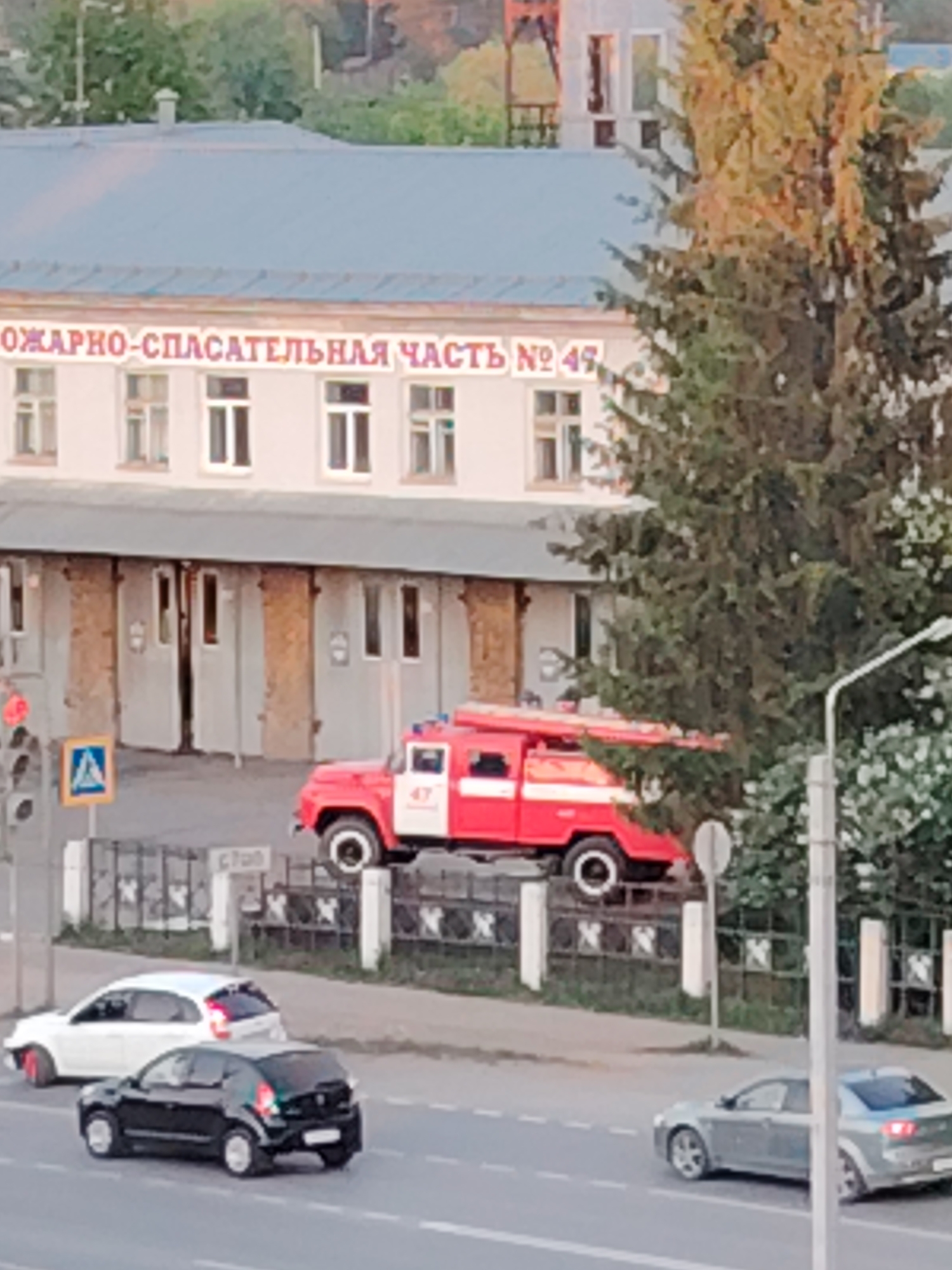 The image shows the Fire and Rescue Unit No. 49 in Chaikovsky, Russia. A red fire engine is parked behind a fence in front of the fire station. Several cars are on the street. A traffic light and street signs are visible. A large evergreen tree stands to the right of the fire station building.