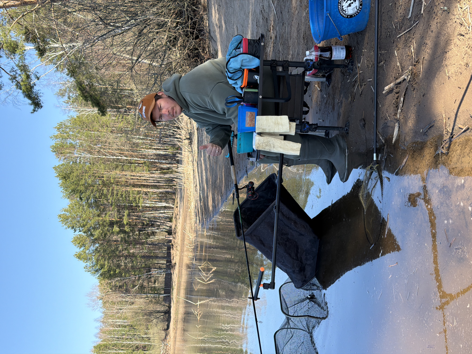The image shows a man standing on a sandy bank next to a body of water, likely a river or lake. He is wearing a green hooded sweatshirt, a brown cap with the word "SAGAME" on it, and green waders. He is giving a thumbs-up gesture.
Next to him is a fishing station set up with various pieces of equipment, including a tackle box, bait containers, a bucket labeled "CHAYKOVSKY FEEDER," bottles, and accessories. A fishing rod is placed nearby, and there are keepnets in the water.
The background features a forest with bare trees and a clear blue sky.
The location is likely near the city of Chaykovsky, Russia, as indicated by the "CHAYKOVSKY FEEDER" label on the bucket. The scene suggests an outdoor recreational activity, specifically fishing.