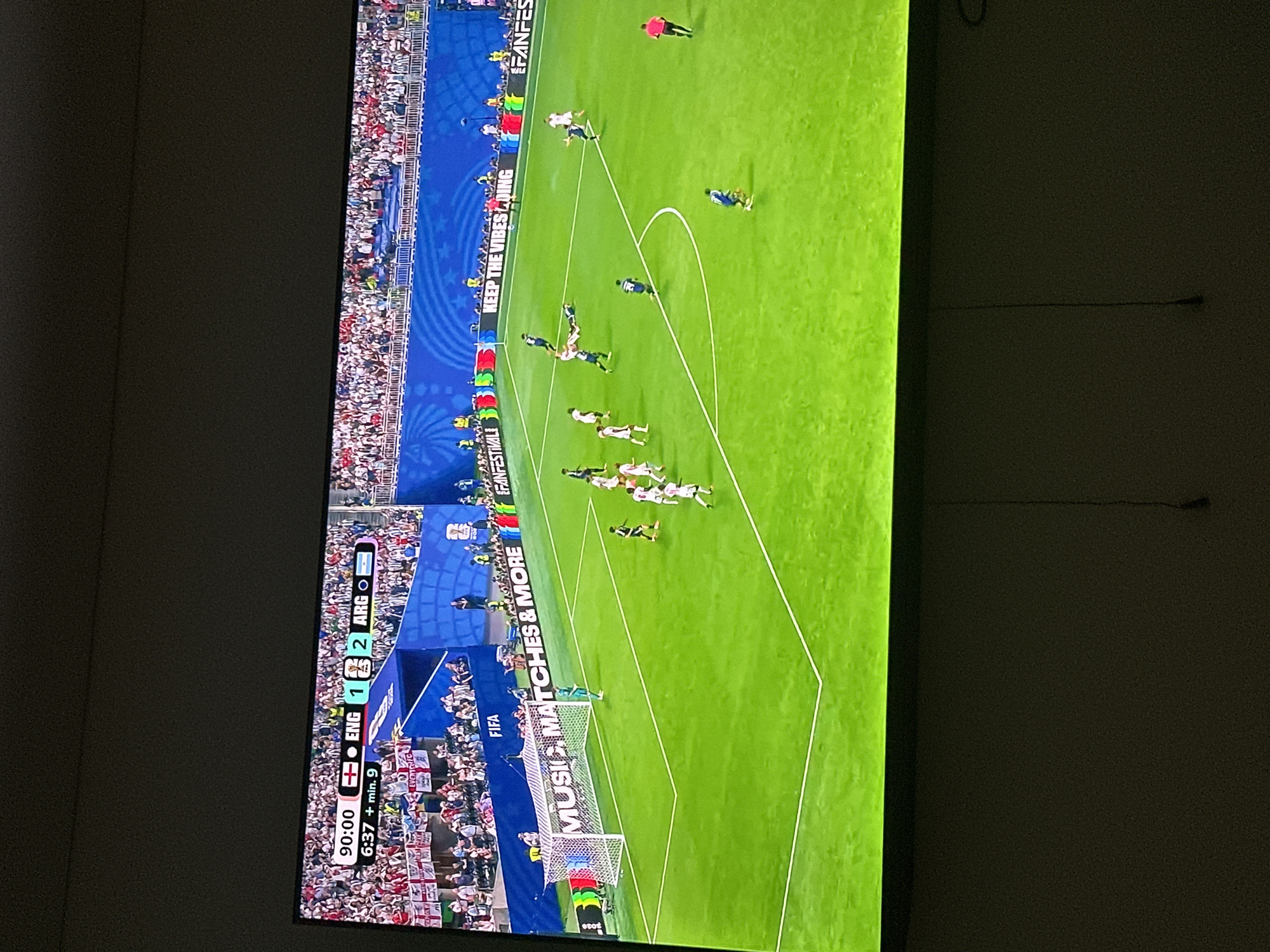 The image shows a screen displaying a soccer match. The score is 1-2, with ARG leading ENG. The time is 90:00 + 9 minutes. The match is taking place in a stadium with a large crowd in the stands. There are players on the field, and a referee. There are also advertisements around the field. The screen is positioned on a wall, and the lower part of the image shows a darkened room with a cable hanging down. This scene is not located in Benimàmet-beniferri, Spain; it is a broadcast of a soccer match.