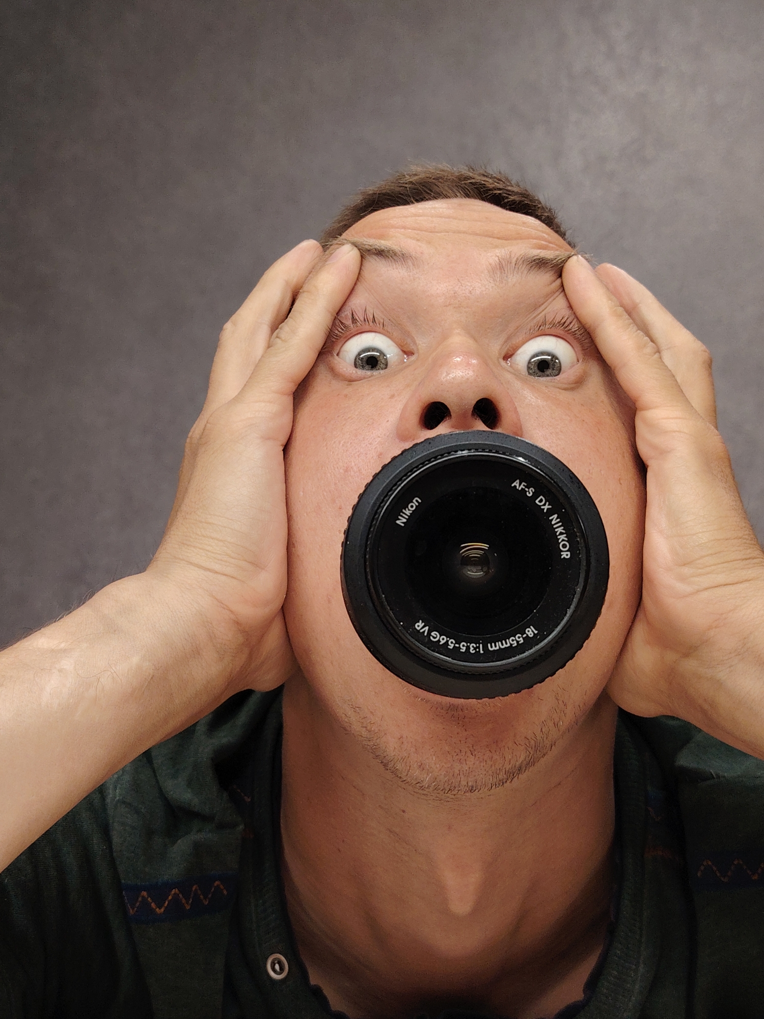 The image shows a close-up, low-angle shot of a person's face with a camera lens in their mouth. The person, appearing to be a young adult male with light skin and brown hair, has wide, surprised eyes and is holding their temples with both hands. The camera lens, a black Nikon AF-S DX NIKKOR 18-55mm 1:3.5-5.6G VR, is positioned in the center of their open mouth. The background is a plain, dark grey wall, indicating an indoor setting. The person is wearing a dark green hooded garment with a patterned trim visible around the collar. There are no identifiable landmarks or contextual clues within the image to suggest a specific location within Chaikovsky, Russia.