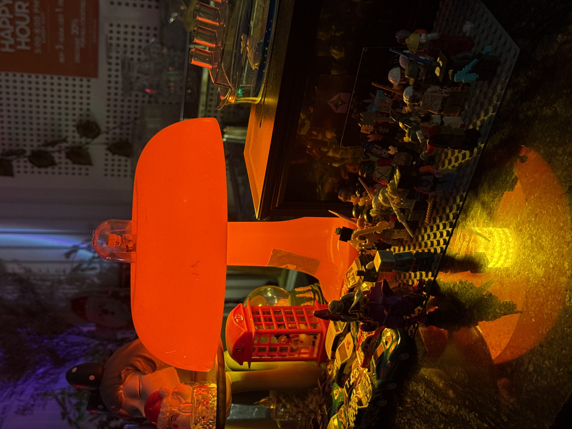 The image depicts a collection of objects on a surface, seemingly a table or display case, with an orange light source dominating the left side. The objects include Lego minifigures arranged on a Lego baseplate, various other plastic toys, and a red telephone booth figurine. Behind these objects, there's a glimpse of an interior space, possibly a store or restaurant, with signage indicating "Happy Hour" and decorative elements like artificial vines. The lighting creates a warm, diffused glow, casting reflections on the surface. No people are visible. The location, Đà Nẵng, Vietnam, is not discernible from the visual information alone.