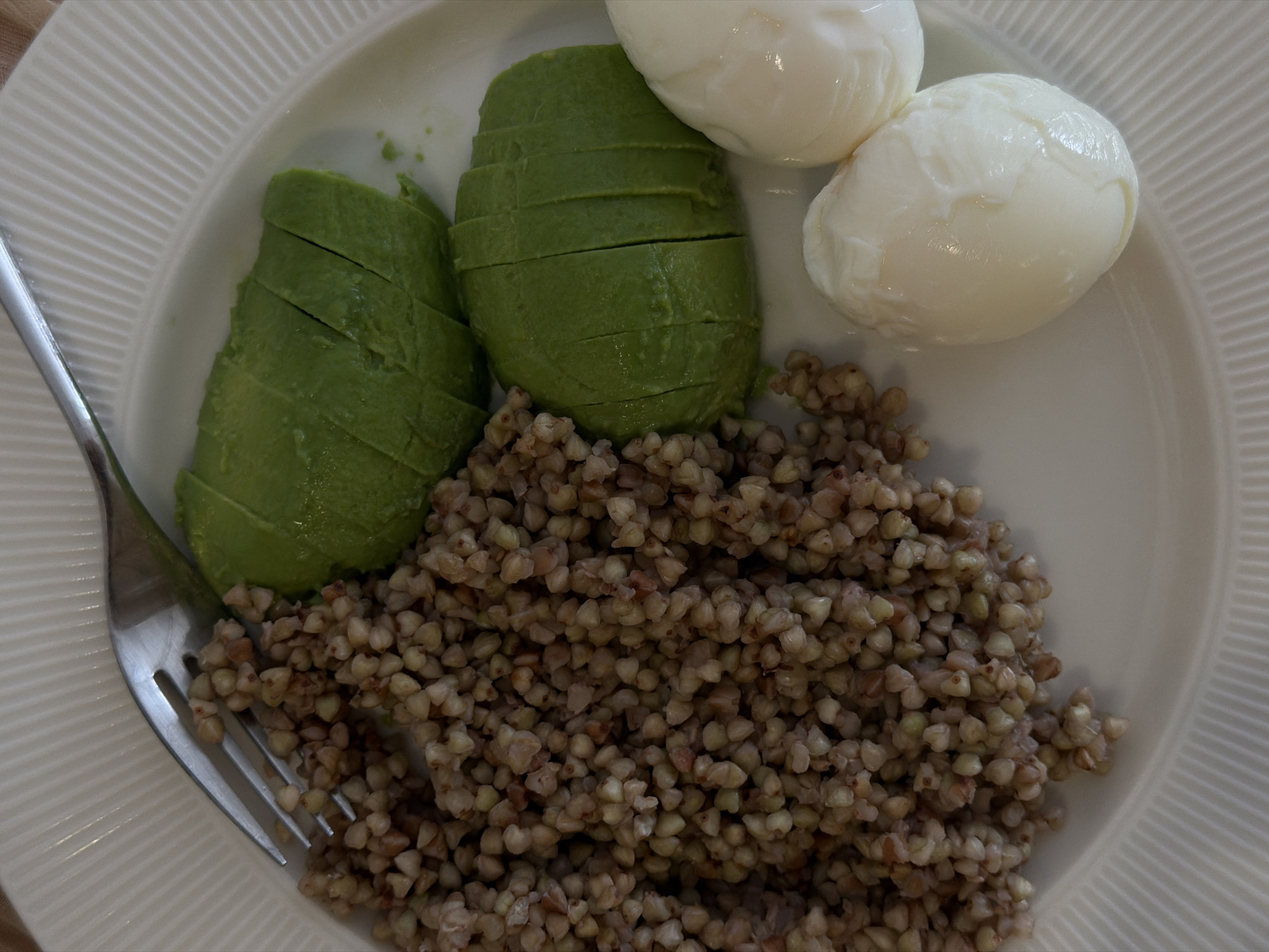 A top-down, medium shot presents a plate of food. The plate, off-white with a ribbed texture, holds a portion of buckwheat, two poached eggs, and sliced avocado. A silver fork rests on the left side of the plate. The lighting appears natural and diffused. No specific location in Budva, Montenegro, is identifiable from the image.