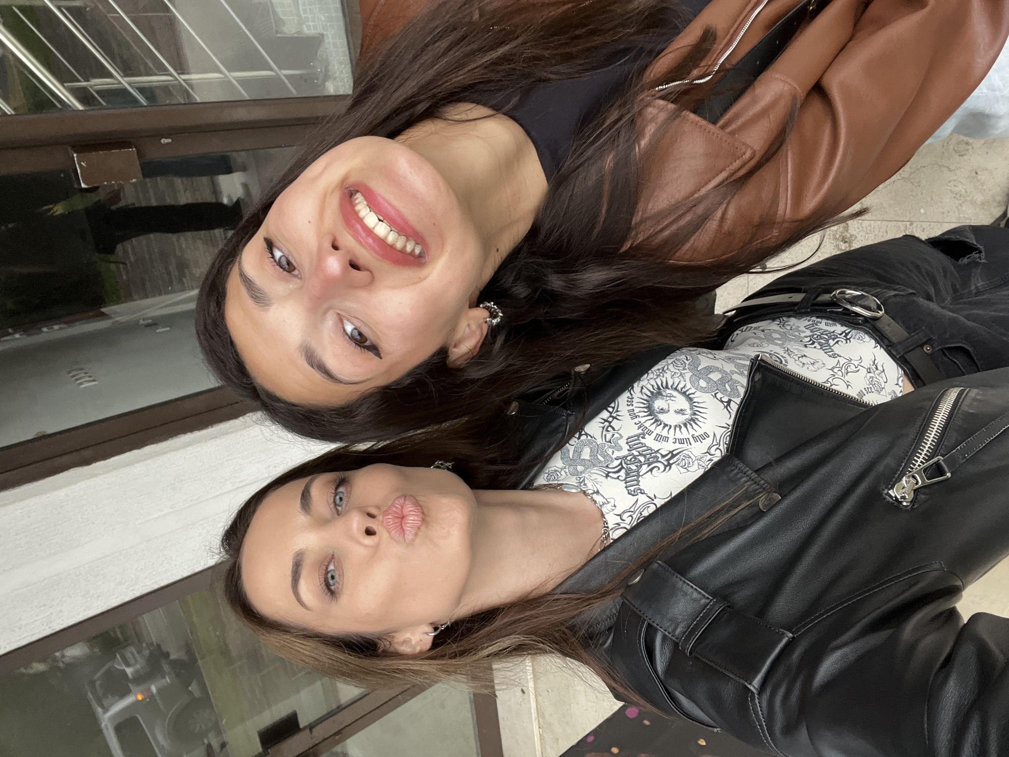 Two women are posing for a photo outdoors. One woman, with dark brown hair and brown eyes, is wearing a brown leather jacket and smiling broadly. The other woman, with light brown hair and blue eyes, is wearing a black leather jacket and puckering her lips. They are standing in front of a building with a window that reflects a vehicle. The location is likely the city of Bar, Montenegro, given the prompt's information.