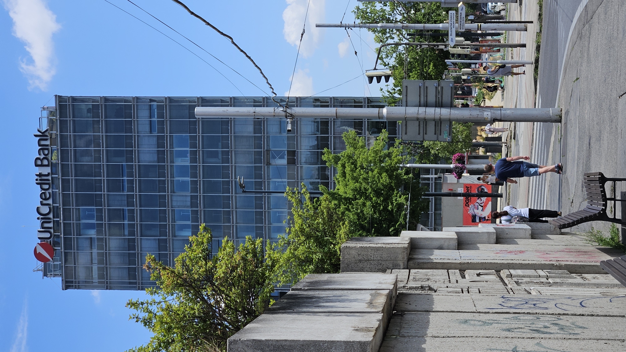 This image captures a street scene in Bratislava, Slovakia, dominated by the UniCredit Bank building, a modern, glass-fronted high-rise. In the foreground, a concrete wall with graffiti runs along the left side of the frame, partially obscuring a wooden bench. Across the street, a pedestrian crossing is visible, with several people walking. A man in a blue t-shirt and shorts is crossing the street, while a woman in a white top and black pants walks away from the camera. In the background, trees with lush green foliage provide a contrast to the urban architecture. Traffic signals and streetlights are also present, indicating a busy intersection. The sky is blue with a few white clouds, suggesting a clear day. The overall atmosphere is one of urban life in a European city.