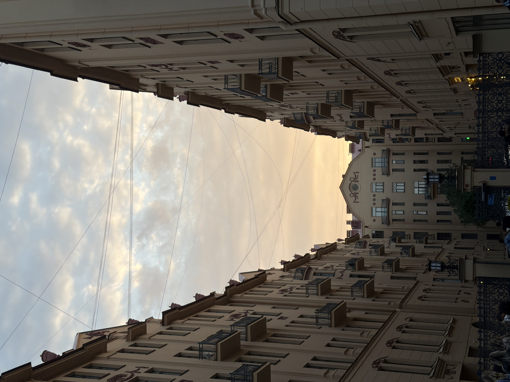 This image shows a street scene in Saint Petersburg, Russia. Two buildings with similar facades and balconies face each other, creating a narrow corridor. The buildings are light beige with repetitive architectural details, including window designs and sculptural elements near the rooftops. Overhead wires crisscross the sky between the buildings. The sky above is filled with clouds and appears to be at sunset or dusk, with warm golden and grey tones. At the end of the street, there is a taller building with a distinctive rounded top. A wrought-iron fence and some blurred figures are visible on the right side of the image.