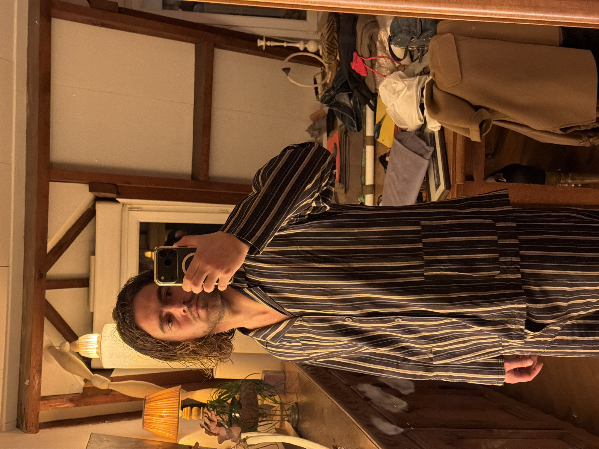 A person is taking a selfie in a mirror. The individual has medium-length, dark hair and a slight beard. They are wearing a striped, button-down shirt. The reflection reveals a room with wooden beams and a window. A lamp with a wooden base and a pleated orange shade is visible, along with a decorative plant in a vase. The mirror reflects clutter, including clothing and other items. The setting is indoors, appearing to be a bedroom or dressing area. There is no explicit information in the image to indicate that the location is Paris, France.