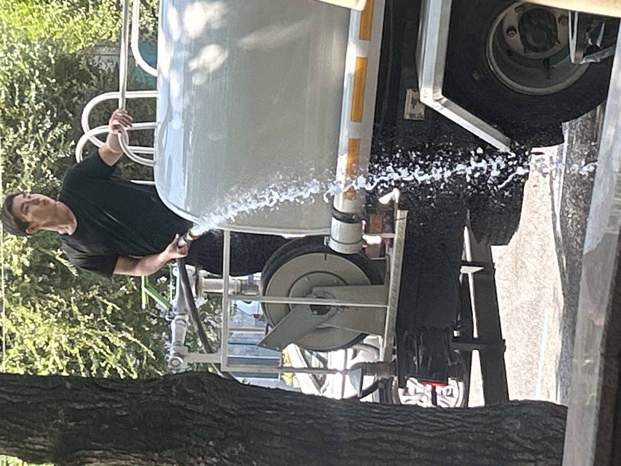 A young man in a black t-shirt and black pants is standing next to a large white water tank truck, holding a hose that is spraying water. The truck appears to be on a city street in Almaty, Kazakhstan, with trees and buildings visible in the background. The water is spraying onto the street and the side of the truck. The man is looking off to the side, away from the camera. The scene is lit by sunlight.