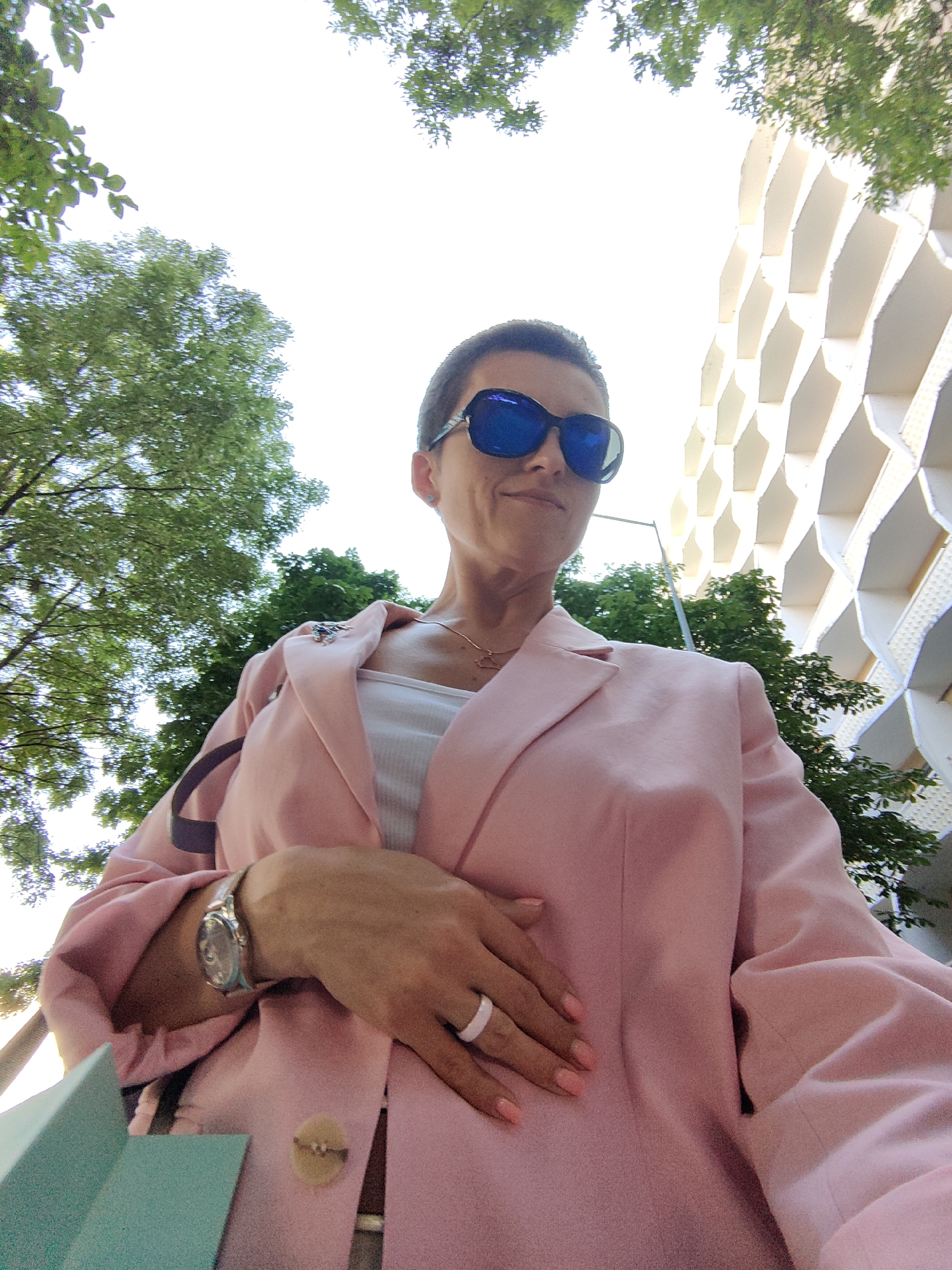 A low-angle, medium shot captures a woman from the chest up, posing outdoors. She wears a pale pink blazer over a white tank top, accessorized with large, blue-tinted sunglasses and a silver watch on her left wrist. A light-colored ring adorns her right ring finger. Her hand rests on her chest, and a small, light-brown button is visible on the blazer near her stomach. A purple strap from a bag is draped over her left shoulder.
In the background, lush green trees with visible leaves frame the top portion of the image. To the right, a modern building with a distinctive honeycomb-like facade, composed of white, hexagonal structures, rises into view. The sky is bright and overcast, suggesting daytime. The scene appears to be set in an urban environment, possibly near a park or a street with contemporary architecture. The lighting suggests it is daytime, with the sun possibly behind the camera, casting soft light on the subject.