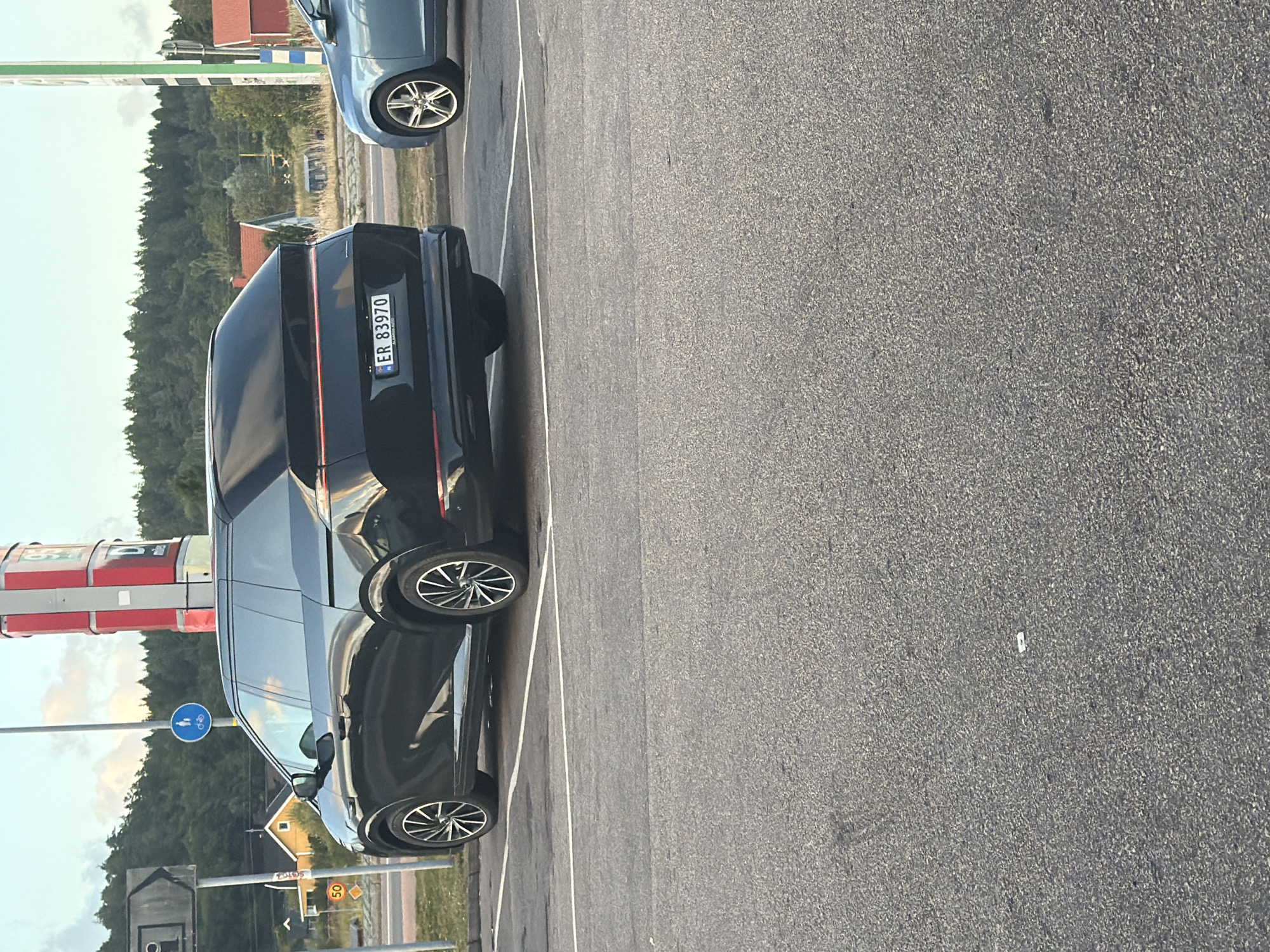 In Hogstorp, Sweden, a black electric car is parked in a lot. The car's license plate reads "ER 83970". A blue car is parked to the right of the black car. In the background, there are trees, houses, and a tall red and white structure that appears to be a gas station sign. A speed limit sign indicating 50 is visible on the left.