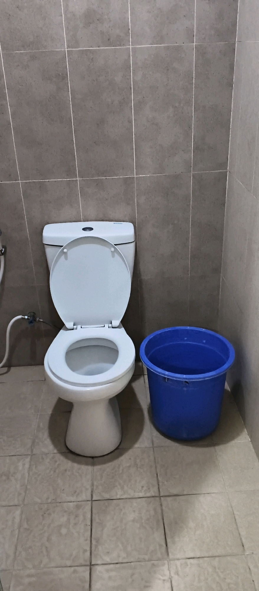 A toilet with the seat up is positioned on a tiled floor. To the right of the toilet, there is a blue plastic bucket. The walls behind the toilet are also tiled. The scene appears to be a bathroom. There are no people present in the image. This scene is located in Tugu, Indonesia.