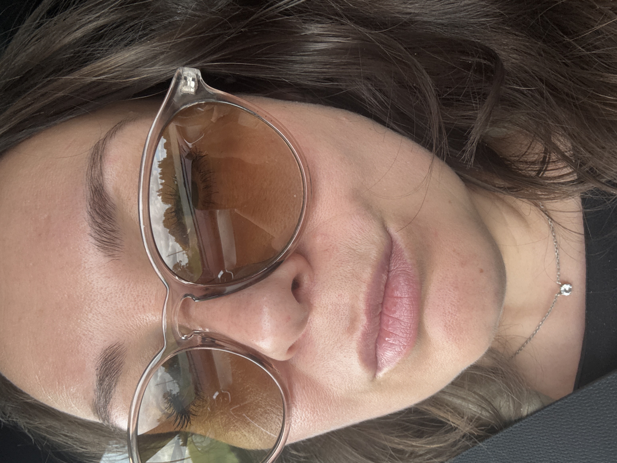 A close-up, waist-up, outdoor selfie of a woman wearing sunglasses and a necklace, looking into the camera. The woman has dark brown hair, fair skin, and is wearing a black top. She is wearing light pink lipstick and her eyebrows are dark brown and arched. The sunglasses have clear frames and amber lenses. The background is blurred, showing trees and a road. The image is likely taken in Krasnoyarsk, Russia, given the reference to the city in the prompt.