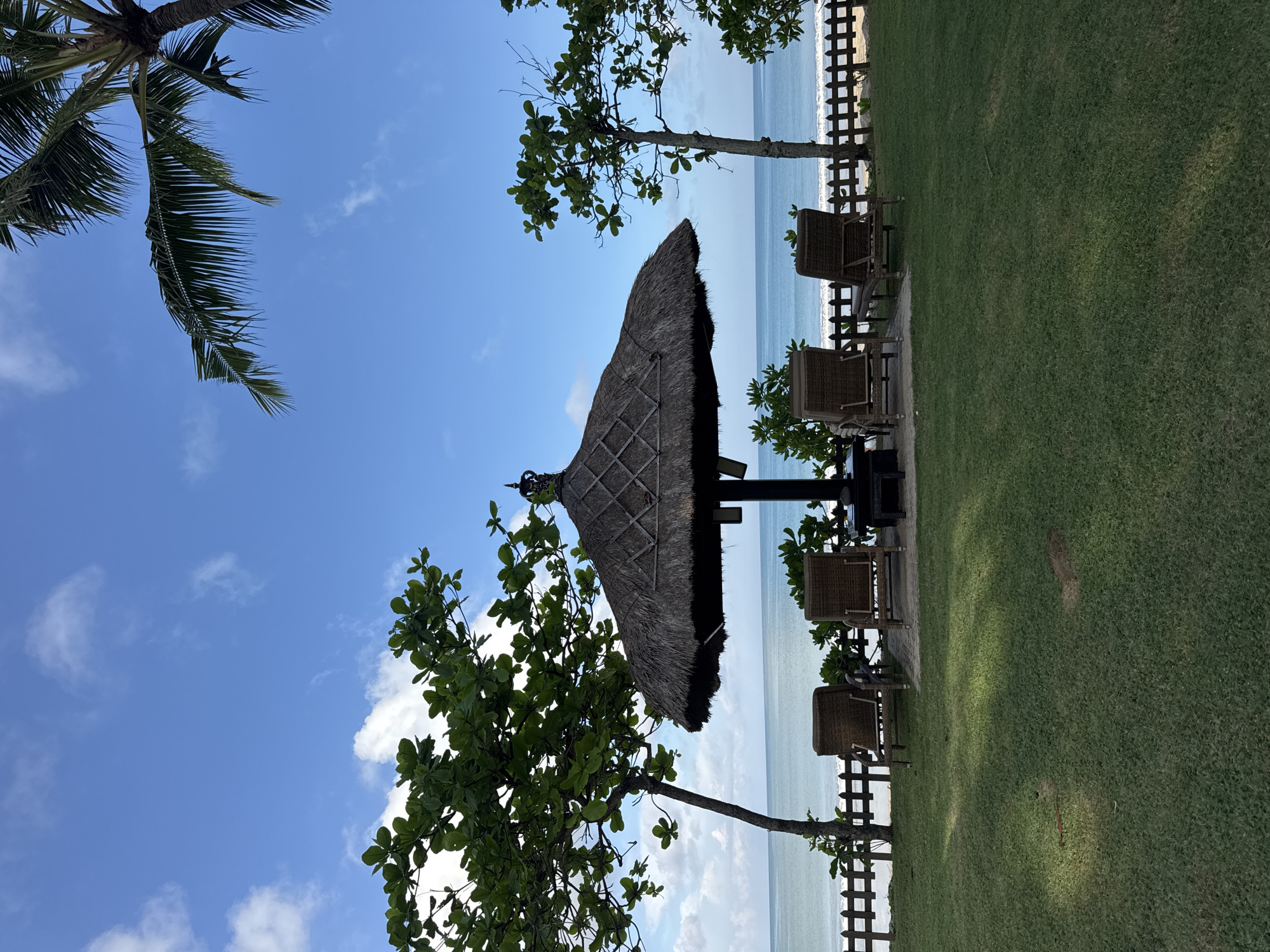 The image depicts a coastal scene, possibly in the city of Jimbaran, Indonesia. A thatched-roof structure stands near a grassy area, likely providing shade. There are four chairs near the roof. A partial view of a palm tree is visible in the top left corner, and other trees are scattered throughout the scene. A wooden fence separates the grassy area from the beach. The sea and sky are visible in the background. The sky is blue with scattered clouds. The image is rotated.