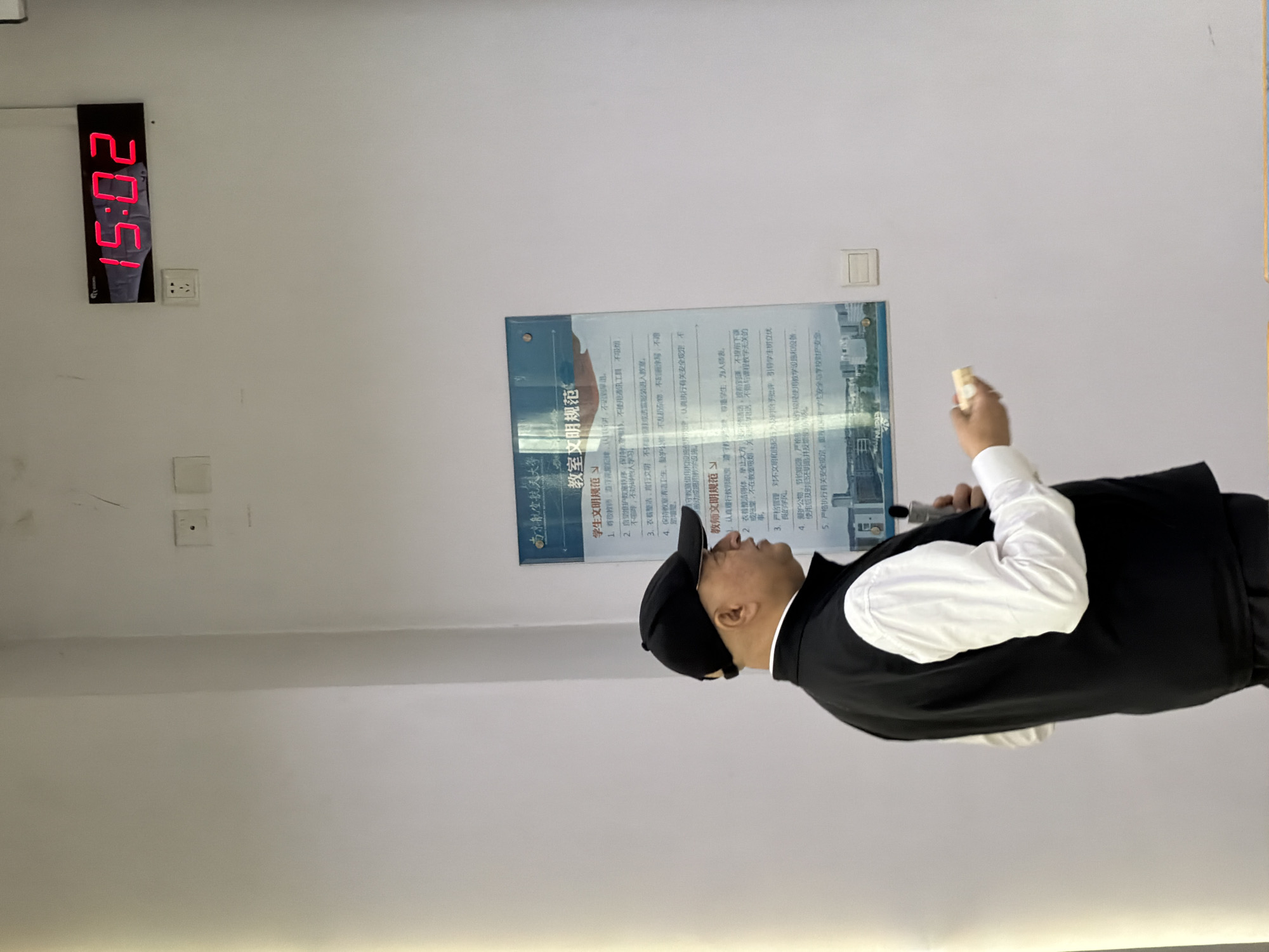 Here is a description of the image:
**Scene**: An indoor shot taken in what appears to be a classroom or lecture hall in Jiangning District, China. The wall is white and unadorned except for a digital clock displaying the time "15:02" (3:02 PM), electrical outlets, and a poster on the wall that reads "Nanjing University of Aeronautics and Astronautics - Classroom Civilized Regulations" in Chinese.
**Person**: A man wearing a black baseball cap, a white long-sleeved shirt, and a black vest stands facing the poster. He holds a small object in his hand (possibly a microphone or pointer) and seems to be in the middle of giving a presentation or addressing a class.
**Poster**: The poster presents rules for students and teachers in Chinese. Key elements visible include sections on "Student Civilized Regulations" and "Teacher Civilized Regulations," listing rules such as "Don't be late or leave early," "Maintain cleanliness," and "Show respect."
**Actions and Interactions**: The man appears to be explaining or lecturing about the regulations on the poster.