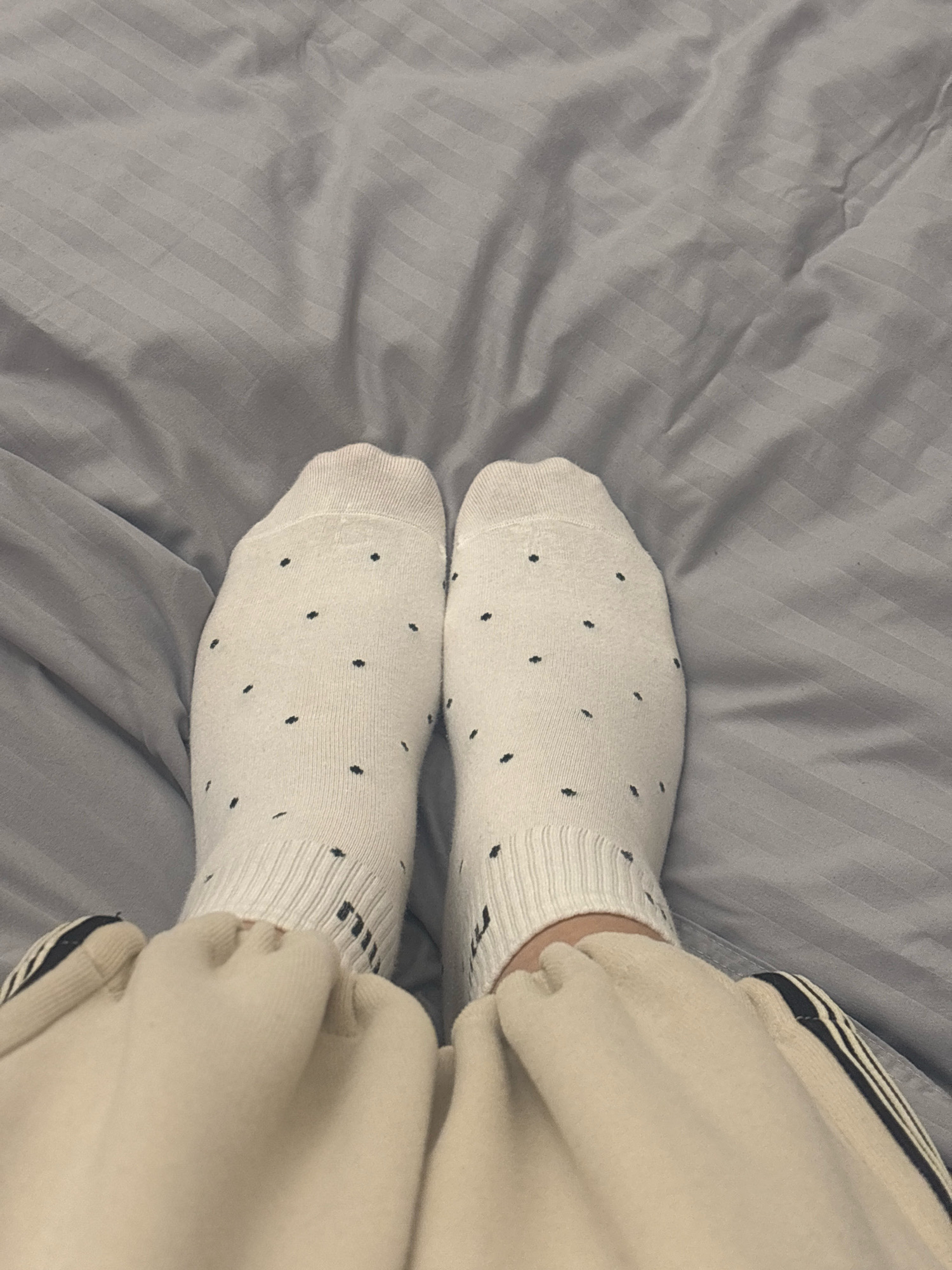 The image shows a pair of white socks with black polka dots on a person's feet, resting on a grey, striped fabric, likely a bedsheet. The socks are ankle-length and appear to be made of a soft, ribbed material. The person is also wearing light-colored pants with a dark stripe detail on the side. The background is a plain, slightly rumpled grey surface with subtle diagonal stripes. There is no visible indication of the location within Getahovit, Armenia, or any urban environment. The scene is static and appears to be indoors.