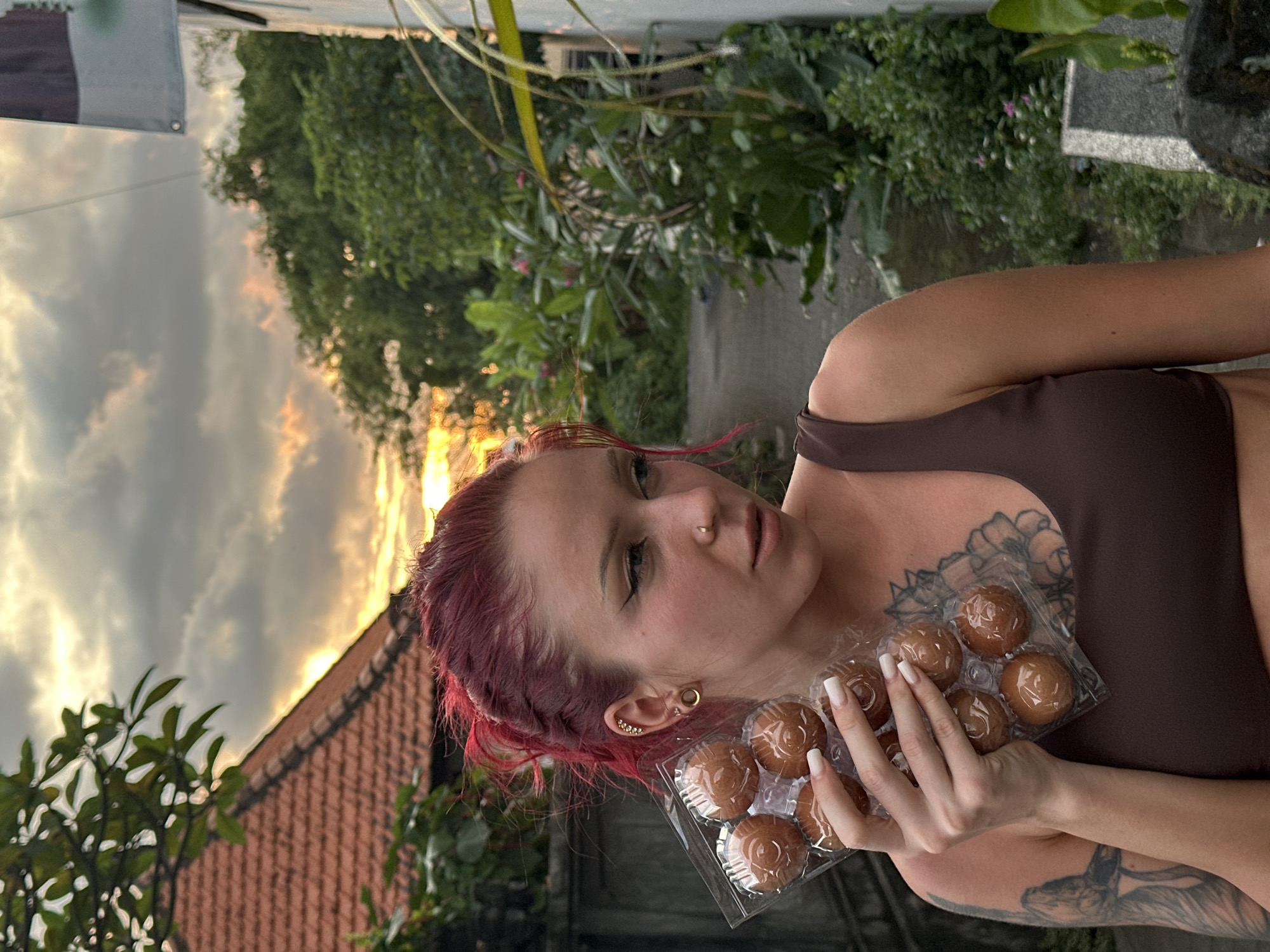 A woman with red hair, a nose ring, tattoos on her chest and arms, and long white fingernails, holds a clear plastic container of nine round, brown pastries. She wears a brown athletic top. The background includes a tiled roof, green vegetation, and a cloudy sky with a glimpse of sunset. The location is likely Dalung, Indonesia.