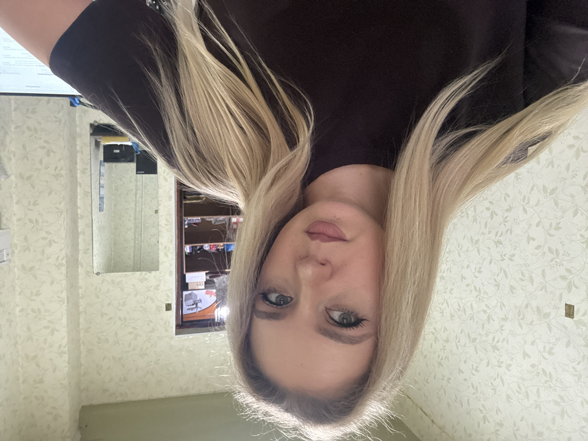 A medium shot, eye-level, selfie-style photograph shows a young Caucasian woman with long, blonde hair, light green eyes, and a neutral expression. She is wearing a dark brown t-shirt. The background consists of a wall with light green, floral-patterned wallpaper, a shelf unit filled with various items, and a mirror. The lighting is even and natural. The location is identified as Unknown, Unknown.