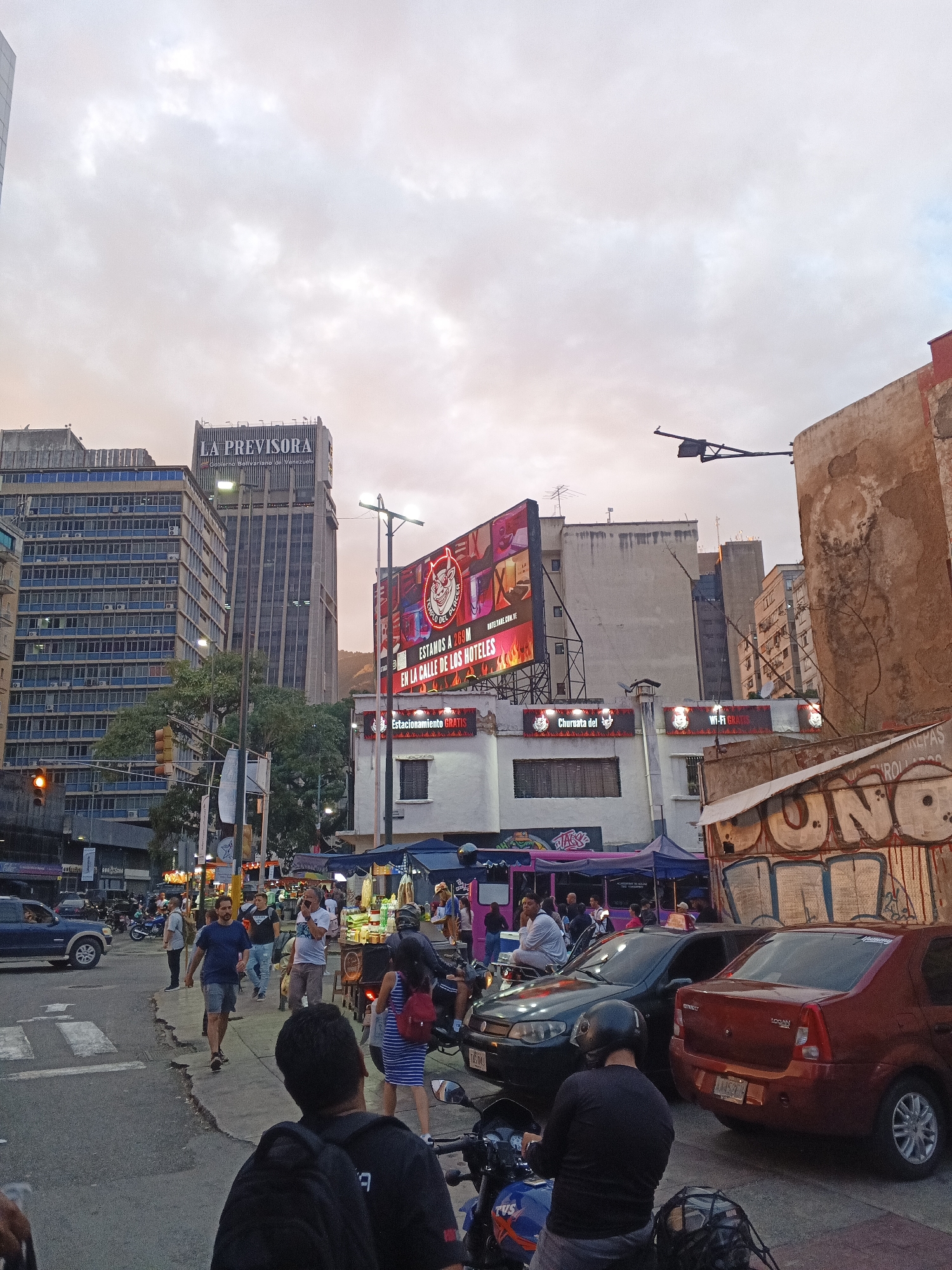 The image captures a street scene in Caracas, Venezuela, likely in a busy urban commercial district. In the background, several tall buildings are visible, including a prominent skyscraper with "LA PREVISORA" signage. A large LED billboard displaying advertisements and text such as "ESTAMOS A 289M EN LA CALLE DE LOS HOTELES" and "WI-FI GRATIS" is mounted on a building. The street level features shops, market stalls with awnings, and a variety of vehicles, including cars and motorcycles. Pedestrians are seen walking on the sidewalks and crossing the street. Traffic signals are present, indicating an intersection. The lighting suggests it is either early morning or late afternoon, with the sky displaying a mix of light clouds and hints of pink and orange. The architecture appears to be a mix of modern and older structures, with some buildings showing signs of wear and graffiti.
