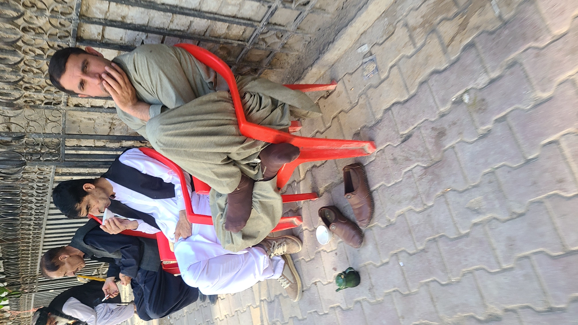 The image shows four men seated on red plastic chairs outdoors, near a sidewalk paved with interlocking concrete blocks. The location appears to be in Quetta City Tehsil, Pakistan.
The man seated in the top right of the frame is wearing a traditional shalwar kameez of muted green and appears pensive. To his left and below him, a man in a white shirt and black vest is drinking from a white cup. To the left is a man holding a pen in his mouth. At the left edge of the frame there is another man in a white shirt and black vest.
Discarded shoes, a small green container, and a cup lid are scattered on the sidewalk near the chairs. A decorative iron fence is visible in the background.