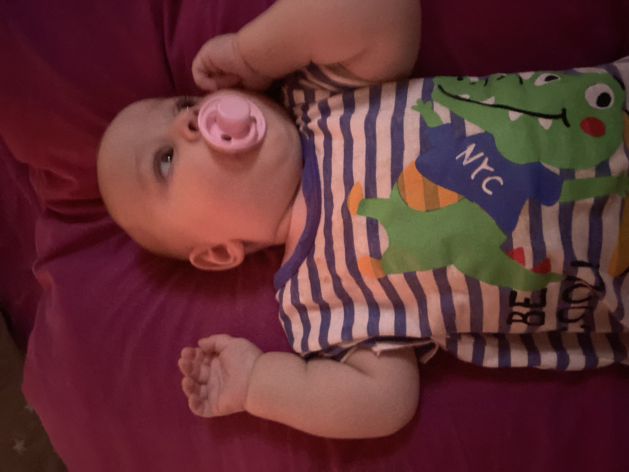 A baby is lying on a dark pink beanbag chair. The baby is wearing a horizontally striped t-shirt with blue and white stripes. The t-shirt has a graphic design featuring a green dinosaur holding a blue sign with "NYC" written in white. The baby has a pink pacifier in its mouth and its hands are near its face. The baby's head is turned to the right, and its eyes are looking in the same direction. The background is the dark pink beanbag chair. The image appears to be taken indoors, with artificial lighting. The location in the city of Otradnaya, Russia, is unknown and cannot be determined from the image.