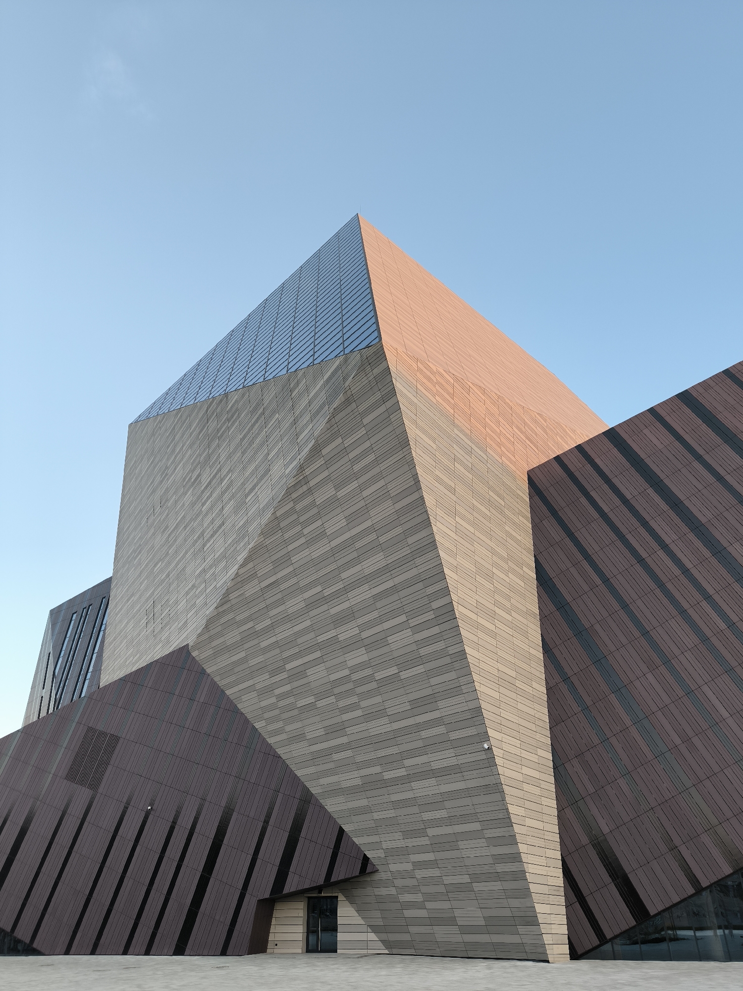 An extreme low-angle outdoor shot shows the faceted exterior of a modern building against a clear blue sky. The building's design features sharp angles and a combination of materials, including what appears to be glass, a sandy-colored textured stone or tile, and a dark brown material with diagonal darker lines. The glass section is at the top of a large angled section, reflecting the sky. Below this, a large, light-colored angled surface dominates the center of the frame, its textured material creating a striped effect. To the right and lower left, large sections of dark brown material with diagonal darker lines create dramatic geometric shapes. A small, dark-colored vent or louvered panel is visible on the dark brown section on the lower left. A security camera is mounted on the light-colored section. The bottom of the image shows a paved area with a recessed entrance featuring double glass doors and a lightly colored frame.
