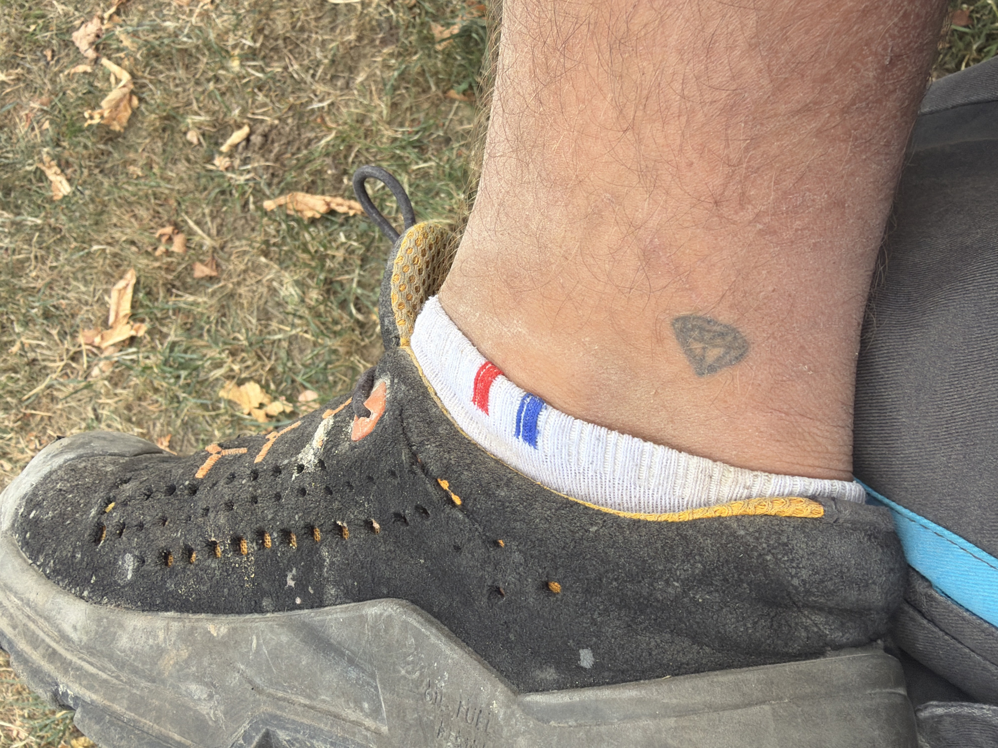The image shows a close-up view of a person's left ankle and part of their shoe, resting on dry grass and fallen leaves. The shoe is dark grey, worn, and appears to be a hiking or outdoor boot with orange accents. A white sock with red and blue stripes is visible above the shoe, around the ankle. On the skin of the ankle, a small, faded black tattoo is present. The tattoo is a geometric shape, possibly a stylized shield or diamond. The background consists of dry grass, dirt, and scattered brown leaves, suggesting an outdoor environment. No specific landmarks are visible to pinpoint a location within Meißenheim, Germany.
