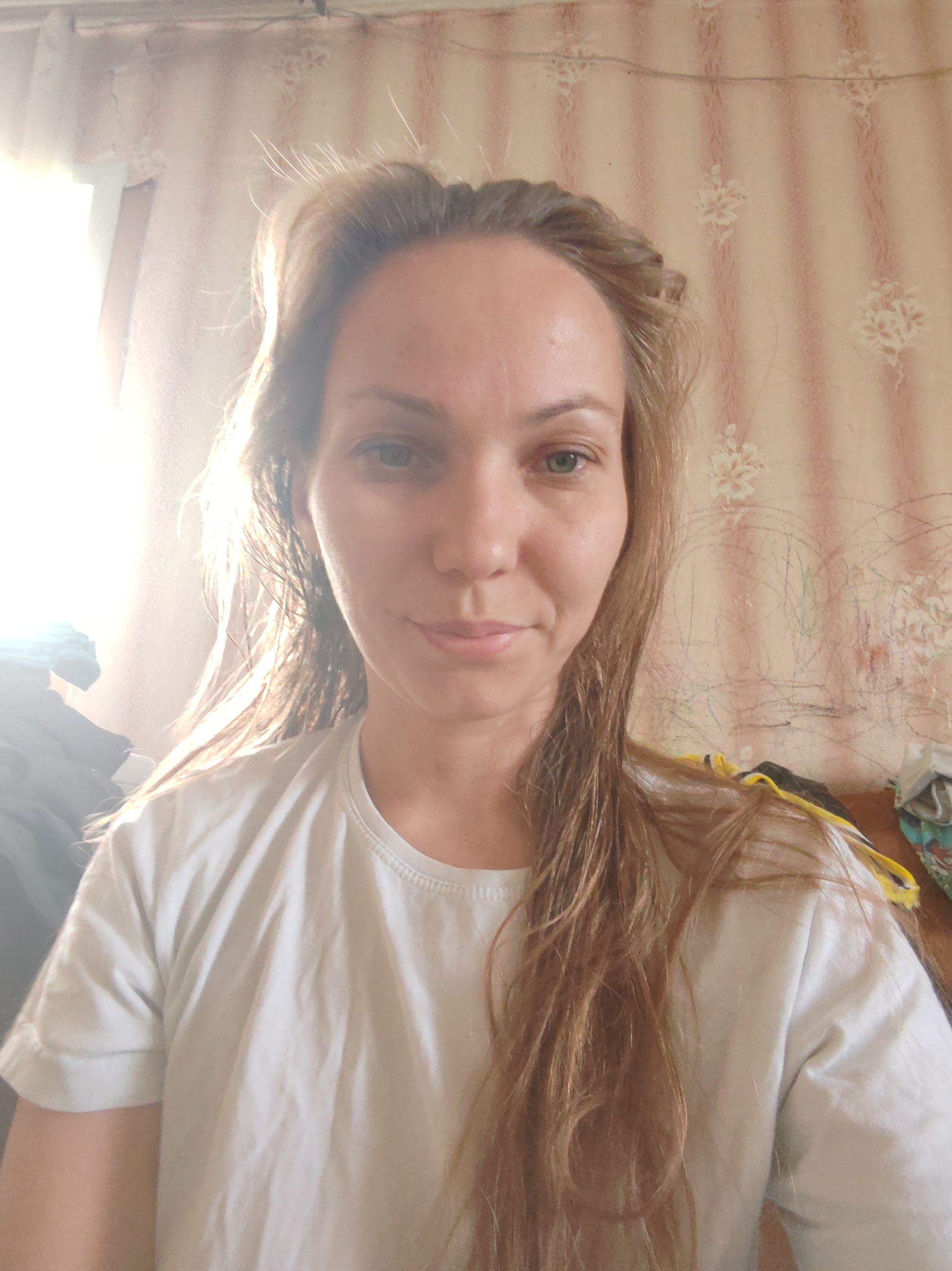 A fair-skinned woman with light brown hair is positioned in the center of the frame. She wears a plain white t-shirt. The background reveals a patterned wallpaper with faint floral designs and vertical stripes. To the left of the frame, there is a window allowing strong sunlight to enter, creating a bright glow. There are some clothes in the left corner of the frame as well. The location could be Чухлэм, Russia, but there are no elements to confirm this information.