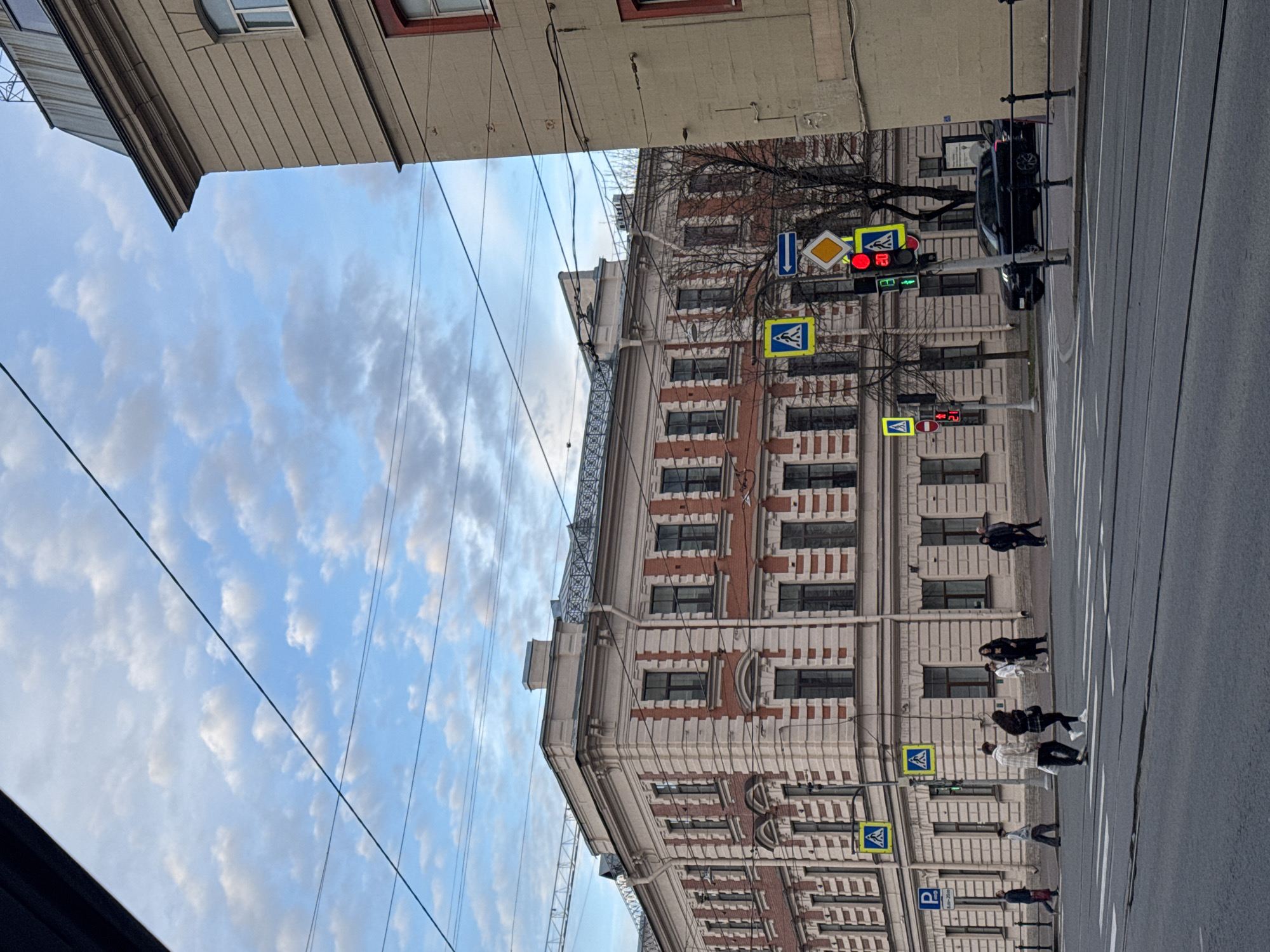 The image shows a street scene in Saint Petersburg, Russia. The buildings are a light color with red brick accents. A row of buildings with multiple windows lines the street. Utility wires are strung across the sky. A traffic light displays red. There are pedestrian crossing signs, and people are walking across the street. A black car is stopped at the traffic light. The sky is blue with white clouds.