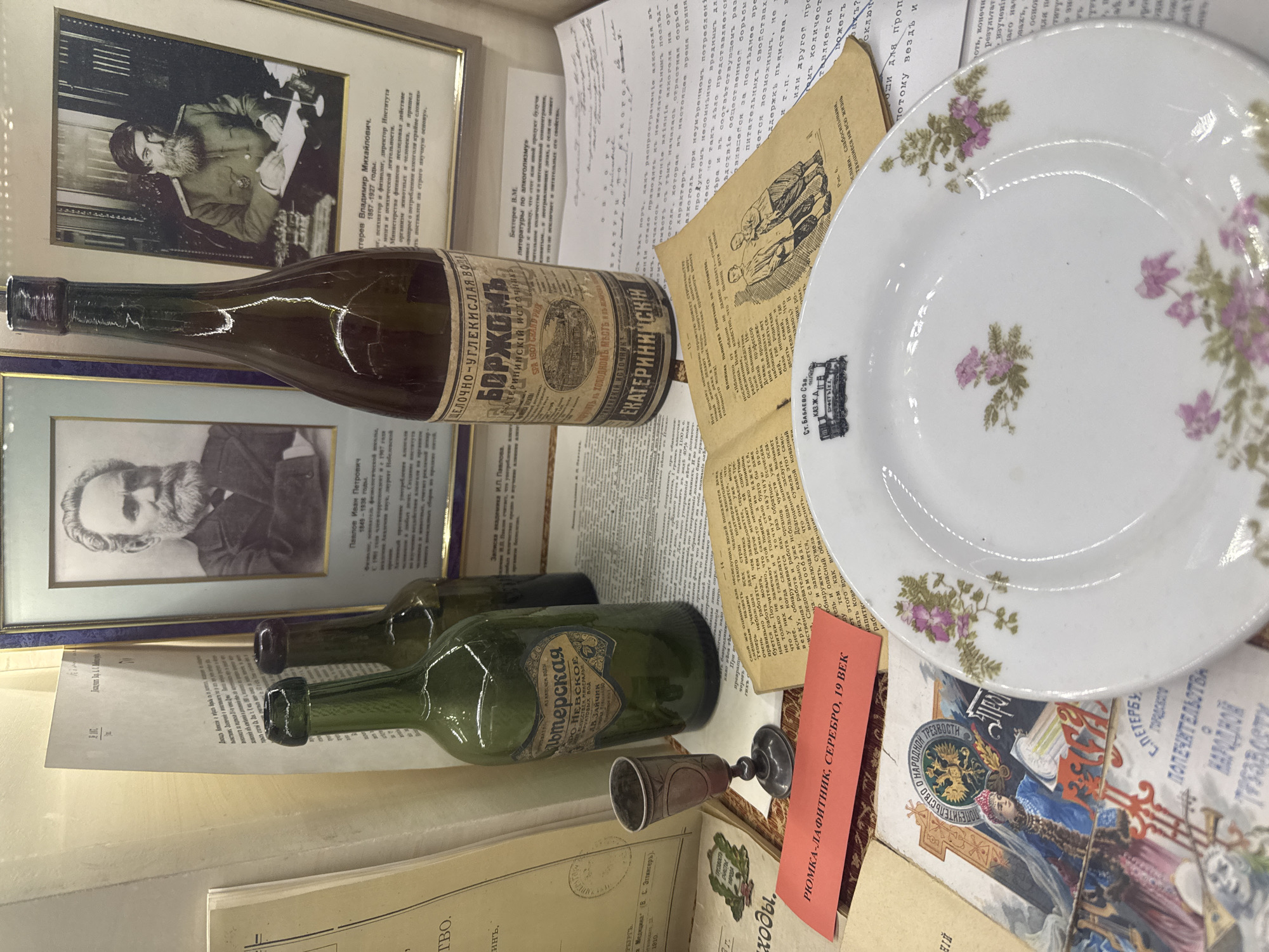 This indoor scene presents a collection of historical items. There are two framed black and white portraits, one of a man with a beard writing at a desk, and another of a man with a beard in a suit. Two vintage bottles, one brown and one green, are placed among documents with Russian text. A white plate with floral designs sits next to an open book with illustrations and a red card with Russian text. A small metal cup rests near the green bottle. The items seem to relate to the history of alcohol consumption and the temperance movement in Russia, evidenced by the bottles labeled "BORJOMI" and "ALTERSKAYA", a leaflet with a title translating to "Society of People's Temperance", and a badge representing a railway stop in Babayevo with a buffet.