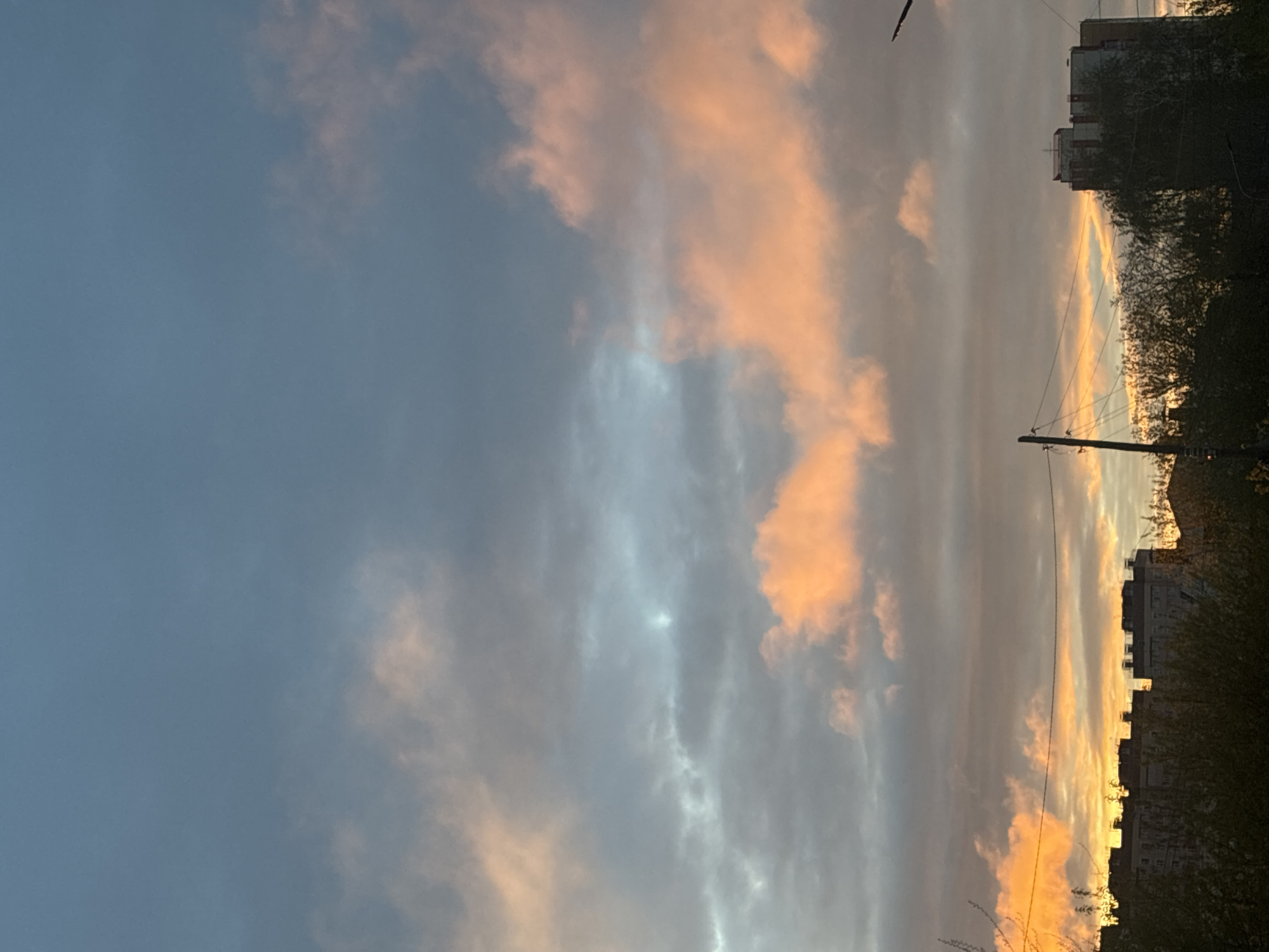 The image shows a dusk or dawn sky in an urban environment. The sky has a range of colors from a dark blue-gray to soft orange and peach hues, suggesting the presence of clouds catching the light of the setting or rising sun. Silhouetted against the sky are buildings and utility poles with power lines. The buildings appear to be multi-story apartment blocks. Trees and shrubbery are also visible in silhouette. The location is Unknown, Unknown.