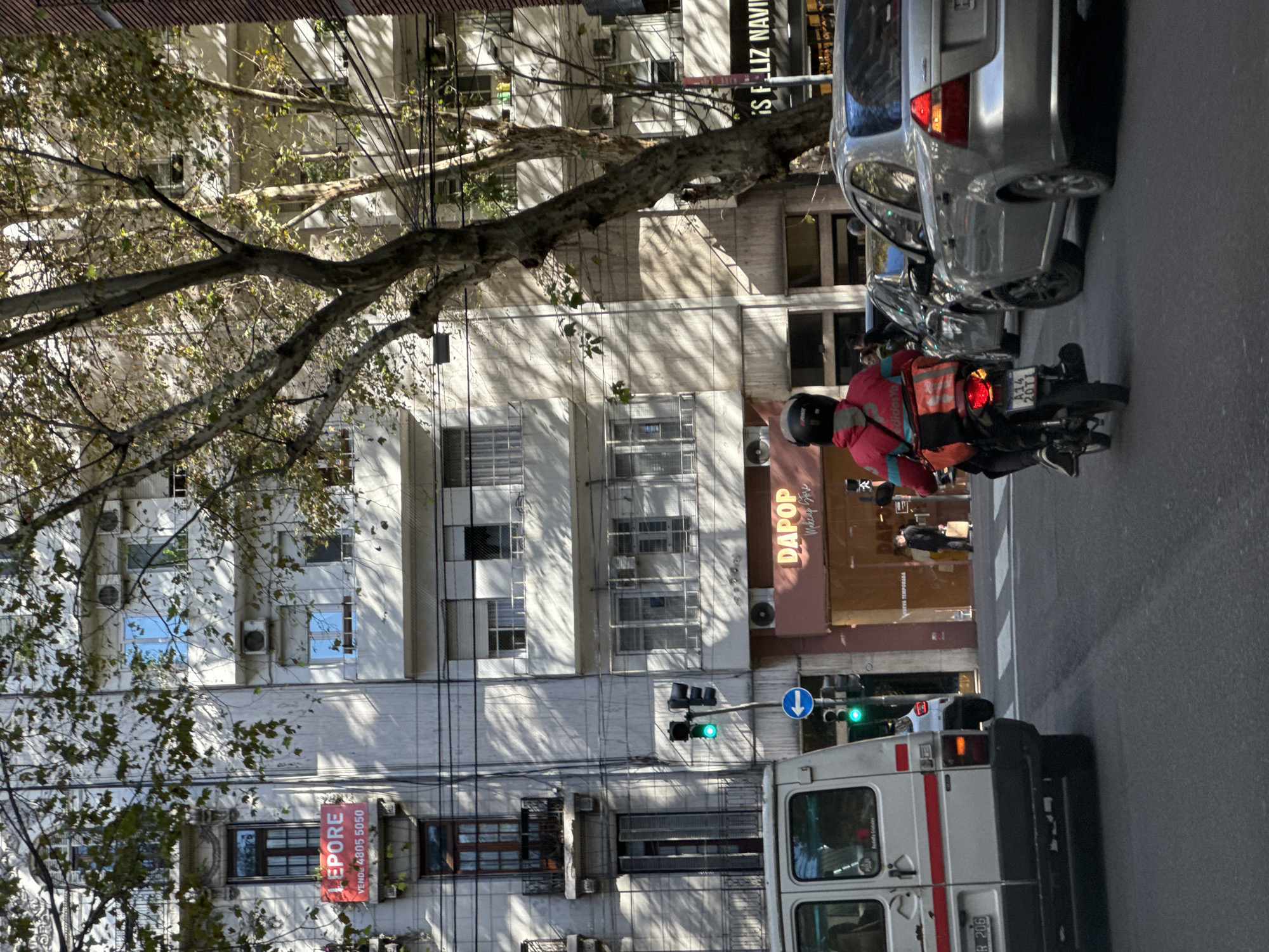 The scene is an urban environment in Buenos Aires, Argentina. The buildings are medium-height, apartment-style structures, many with visible air conditioning units and window security bars. A large tree with bare branches is prominent in the foreground. A motorcycle rider wearing a red jacket and helmet is stopped near a silver car. A white van with red stripes is partially visible. A sign reads "DAPOP Makeup Store." Another sign says "LEPORE VENDE 4805 5050." A traffic light indicates green. A sign also reads "ES FELIZ NAVI." A person is walking on the sidewalk in front of the store.