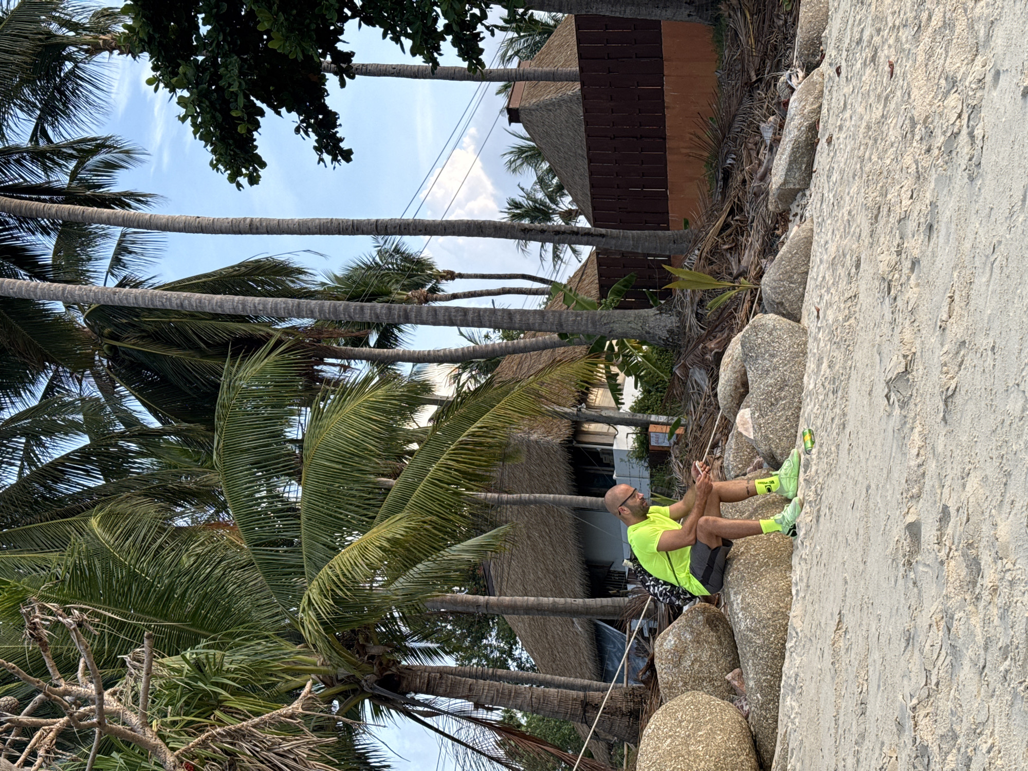 A man with glasses and a shaved head, wearing a neon yellow t-shirt, gray shorts, and bright green athletic shoes, sits on large rocks on a sandy beach. He is looking down at his phone. A backpack with a black and white pattern is on his back. The background shows palm trees, a building with a thatched roof, and a brown wooden fence. The scene appears to be on the island of Ko Samui, Thailand, a popular tourist destination known for its beaches and tropical environment. The lighting suggests it is late afternoon or early evening.