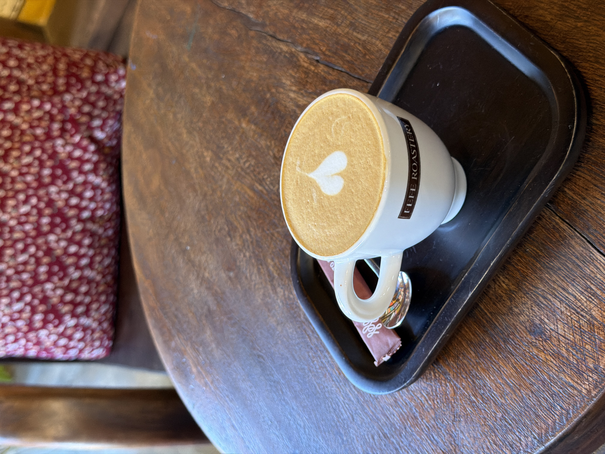 A close-up, top-down shot shows a white ceramic coffee cup on a dark square tray, placed on a rustic wooden table. The cup contains a cappuccino with latte art in the shape of a heart. A small pink packet of sugar and a silver spoon rest on the tray beside the cup. The cup has a black label with the text "FEFE ROASTERY" printed in white. In the blurred background, a plush seat with a red and white patterned cushion is visible. The scene appears to be set in a cafe in Hội An Ward, Vietnam.