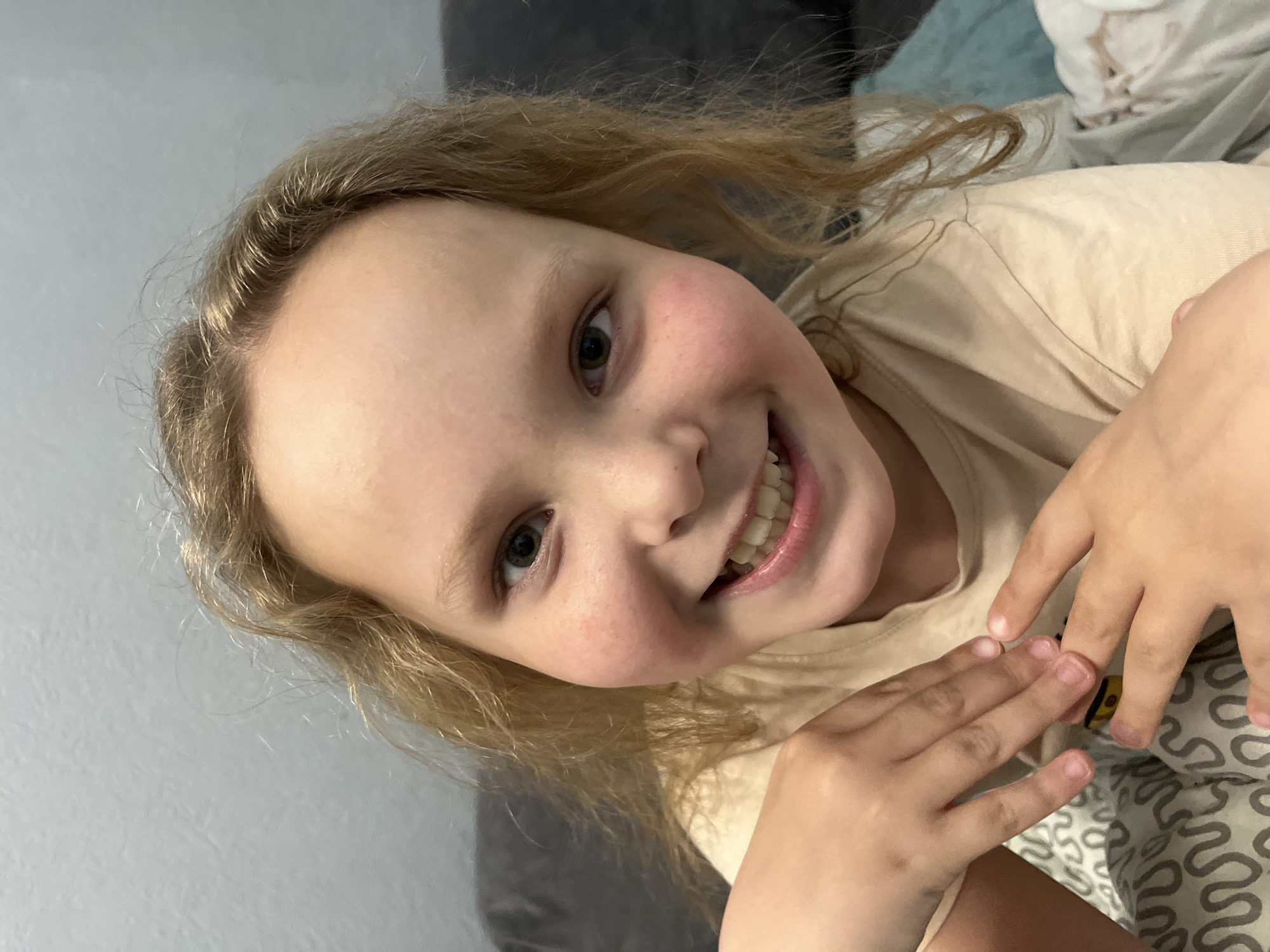 A young, fair-skinned child with blonde, wavy hair and rosy cheeks smiles broadly at the camera. She wears a light beige t-shirt. Her hands are raised in front of her, with her fingers slightly curled. The background includes a gray sofa or wall to the left and a patterned blanket or bedspread to the right. The location is unknown, and there is no evidence to indicate the city of Izhevsk, Russia.